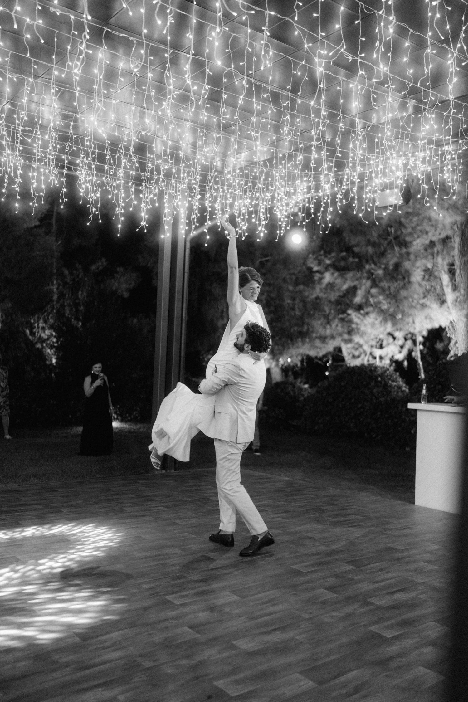 Groom lifts bride during first dance under fairy lights at outdoor wedding reception in black and white photo.