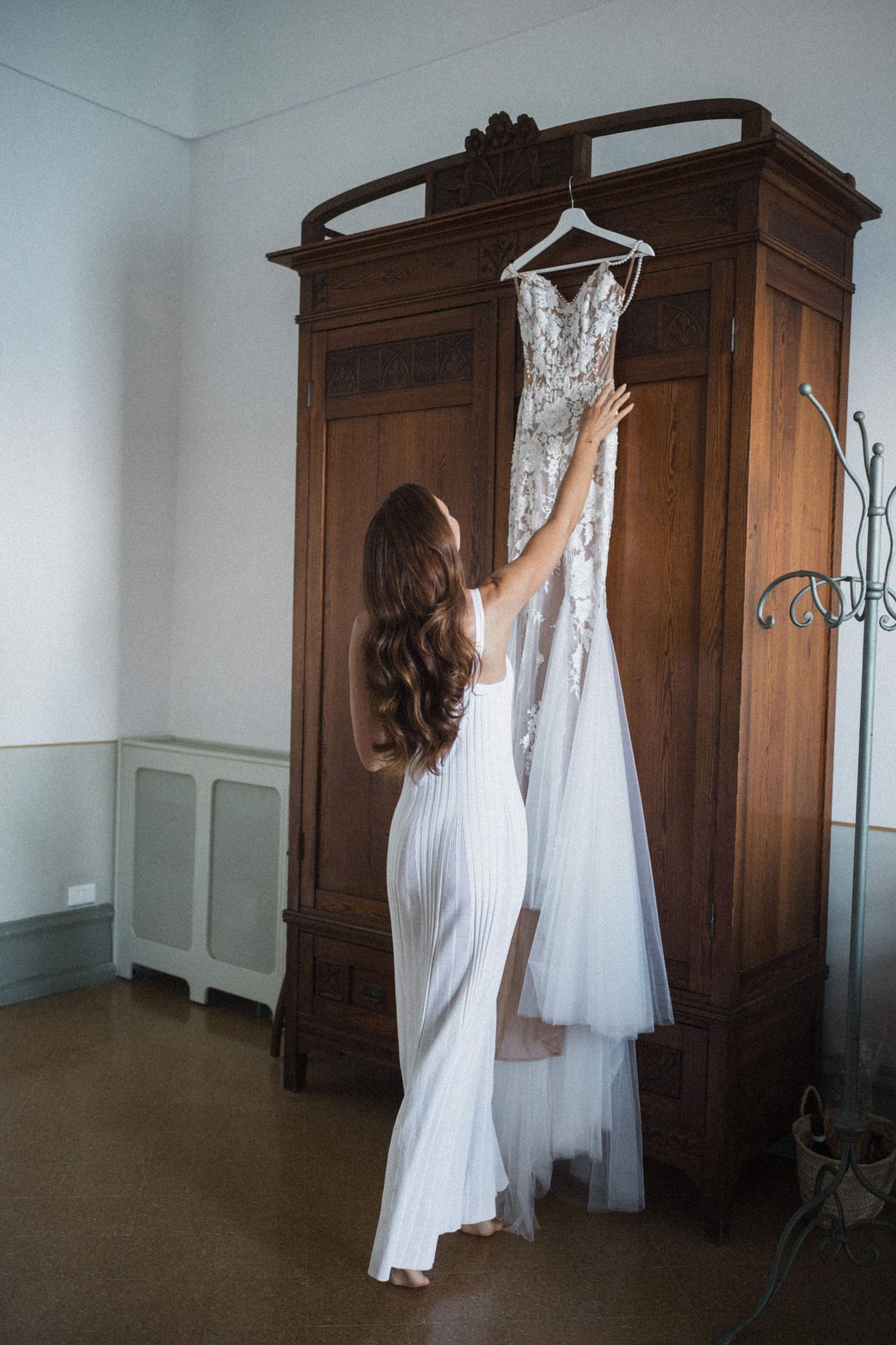 Bride in white slip dress reaching for lace wedding gown hanging on ornate wooden armoire before ceremony.