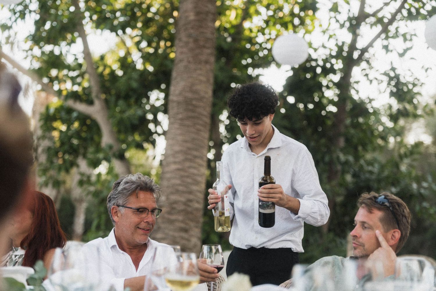 A waiter pours wine for guests seated at an elegant outdoor wedding reception dinner.