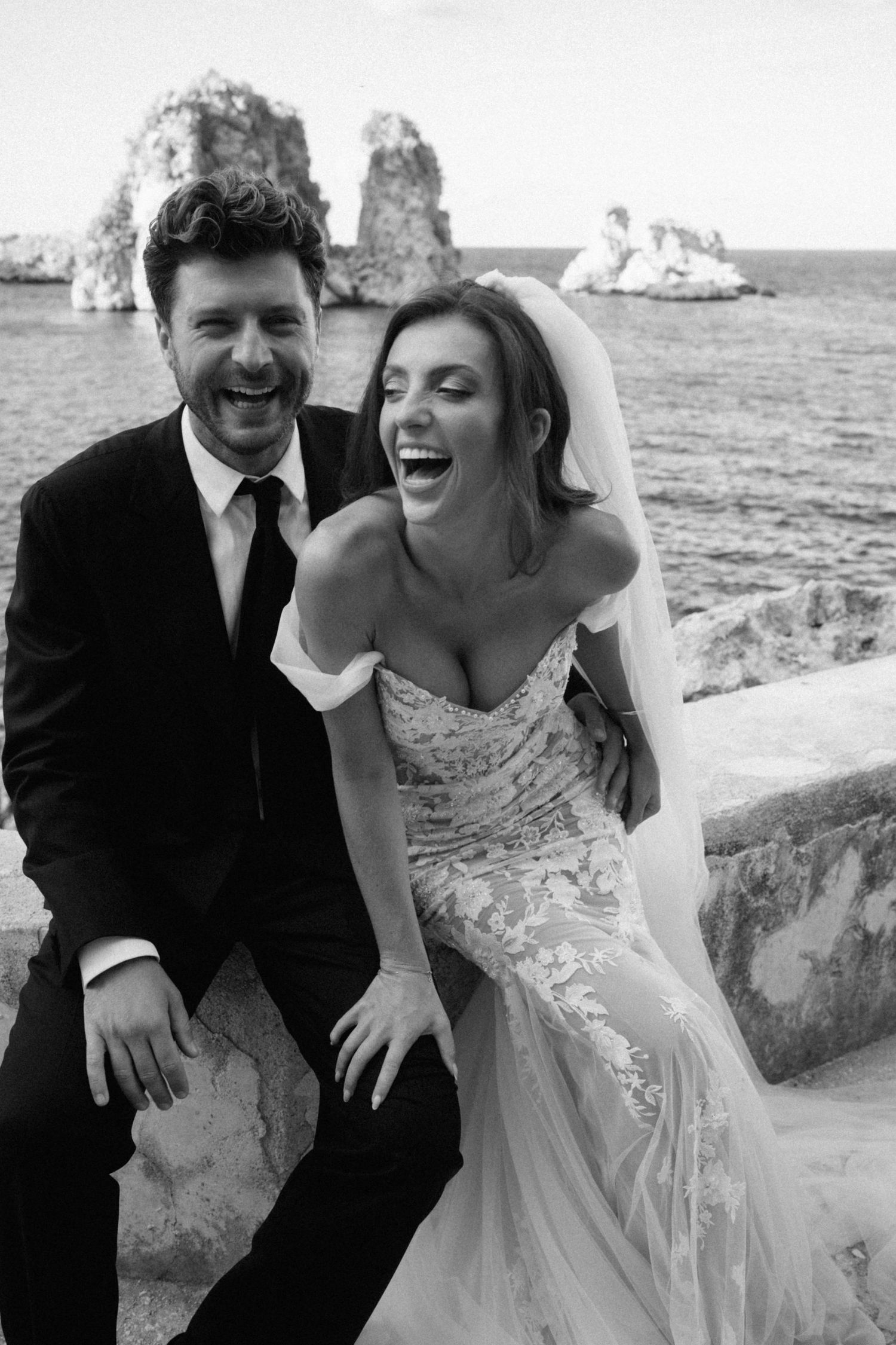 Black and white photo of laughing bride and groom sitting together with rocky ocean coastline behind them.