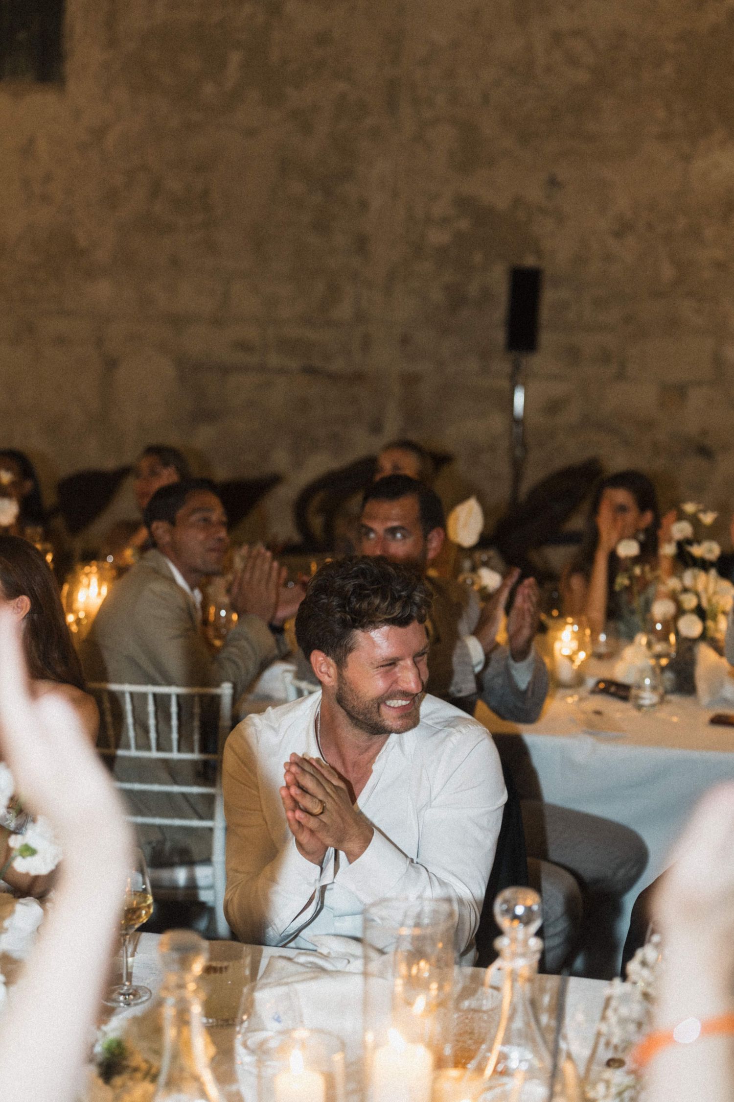 Wedding guest in white shirt claps and smiles during reception dinner at candlelit table.