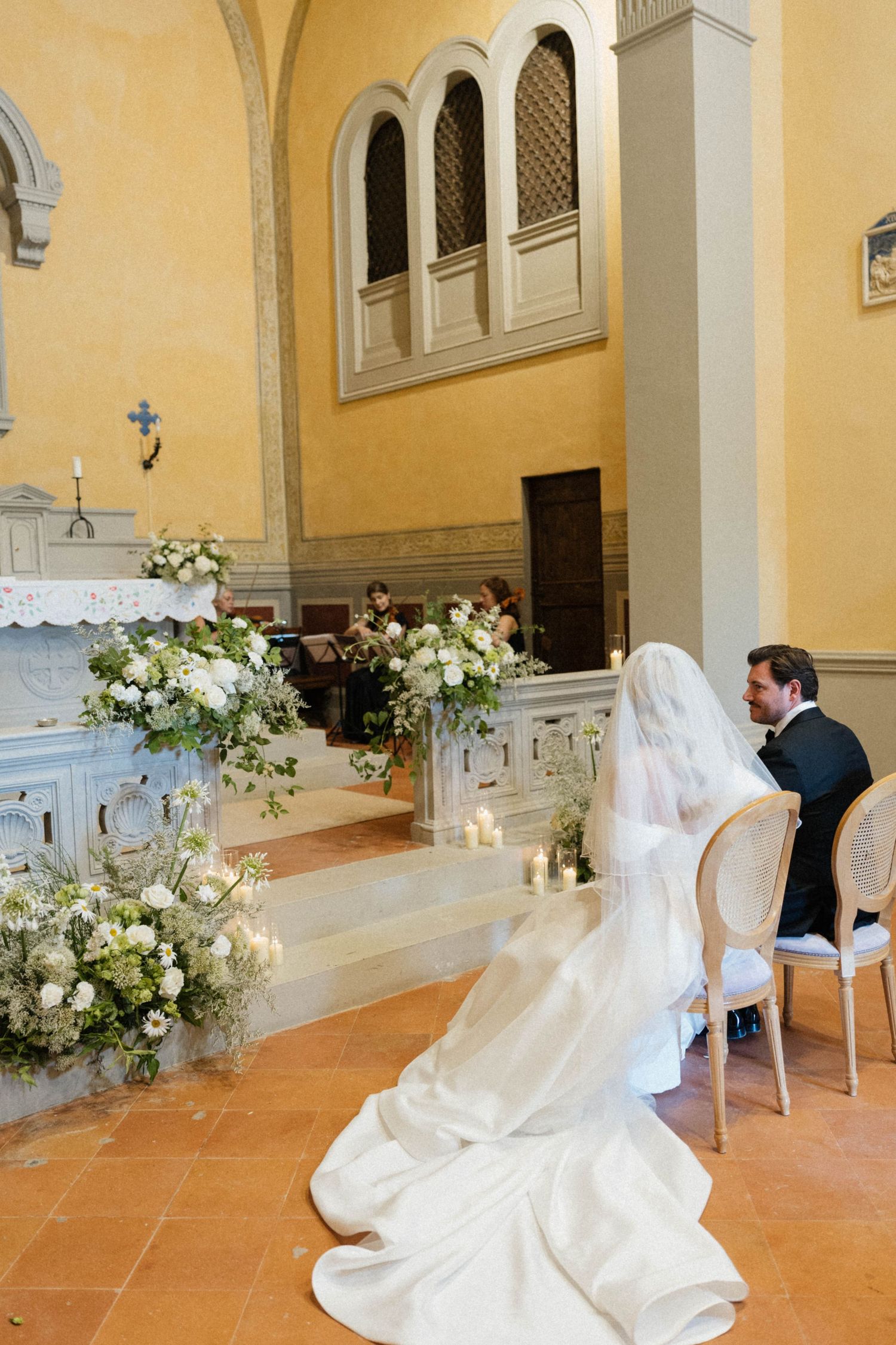 Bride in cathedral veil and groom seated at altar adorned with white floral arrangements.