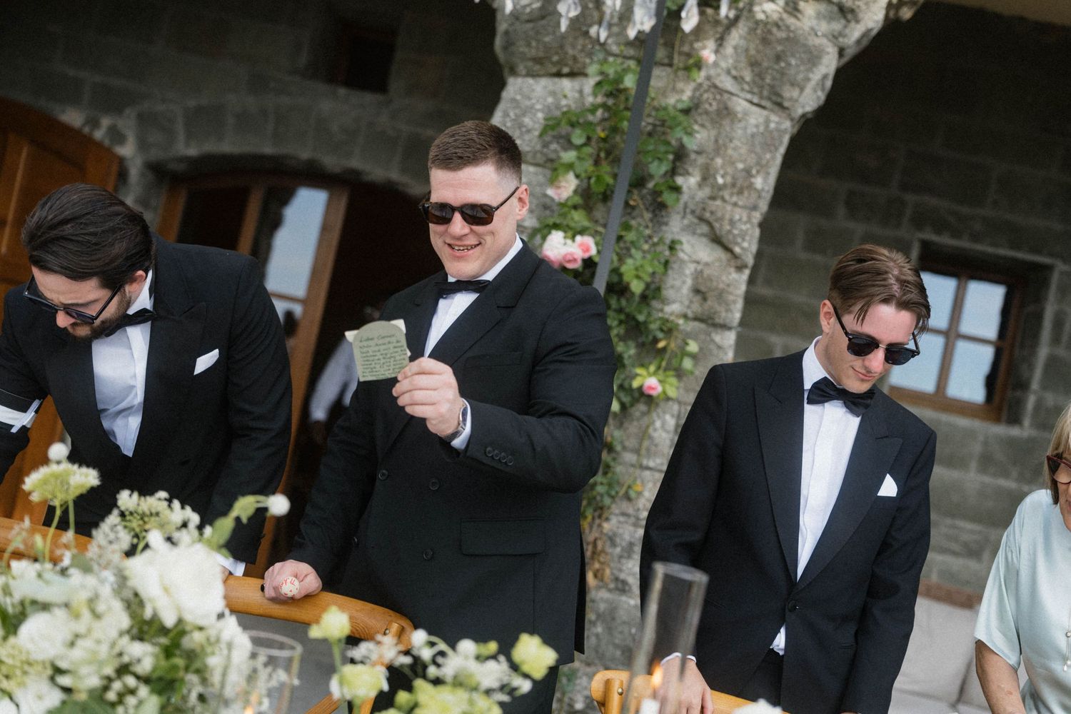 Three groomsmen in black tuxedos with sunglasses stand at floral wedding reception table outdoors.