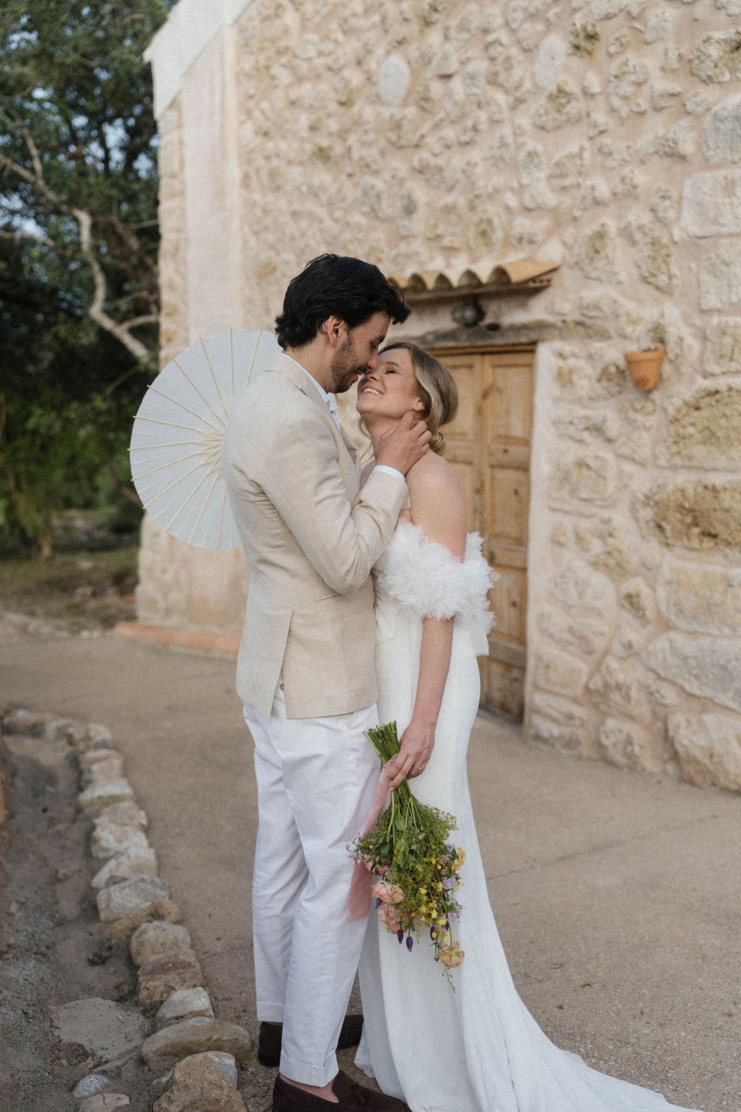 Newlyweds embracing tenderly outside historic stone venue bride holding colorful wildflower bouquet.