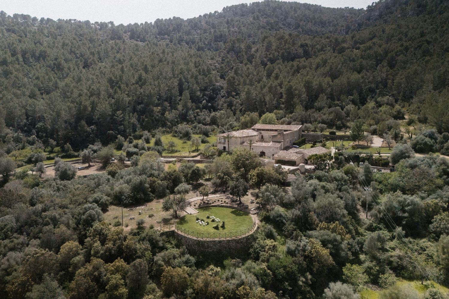 Aerial view of a rustic estate surrounded by lush green forest and olive trees in a serene valley.