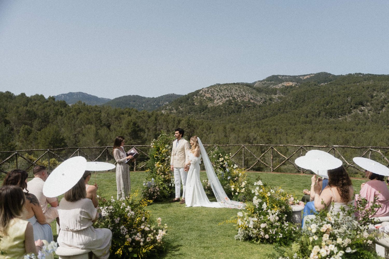Bride and groom exchange vows at outdoor ceremony with mountain views, floral arrangements, and guests holding parasols.