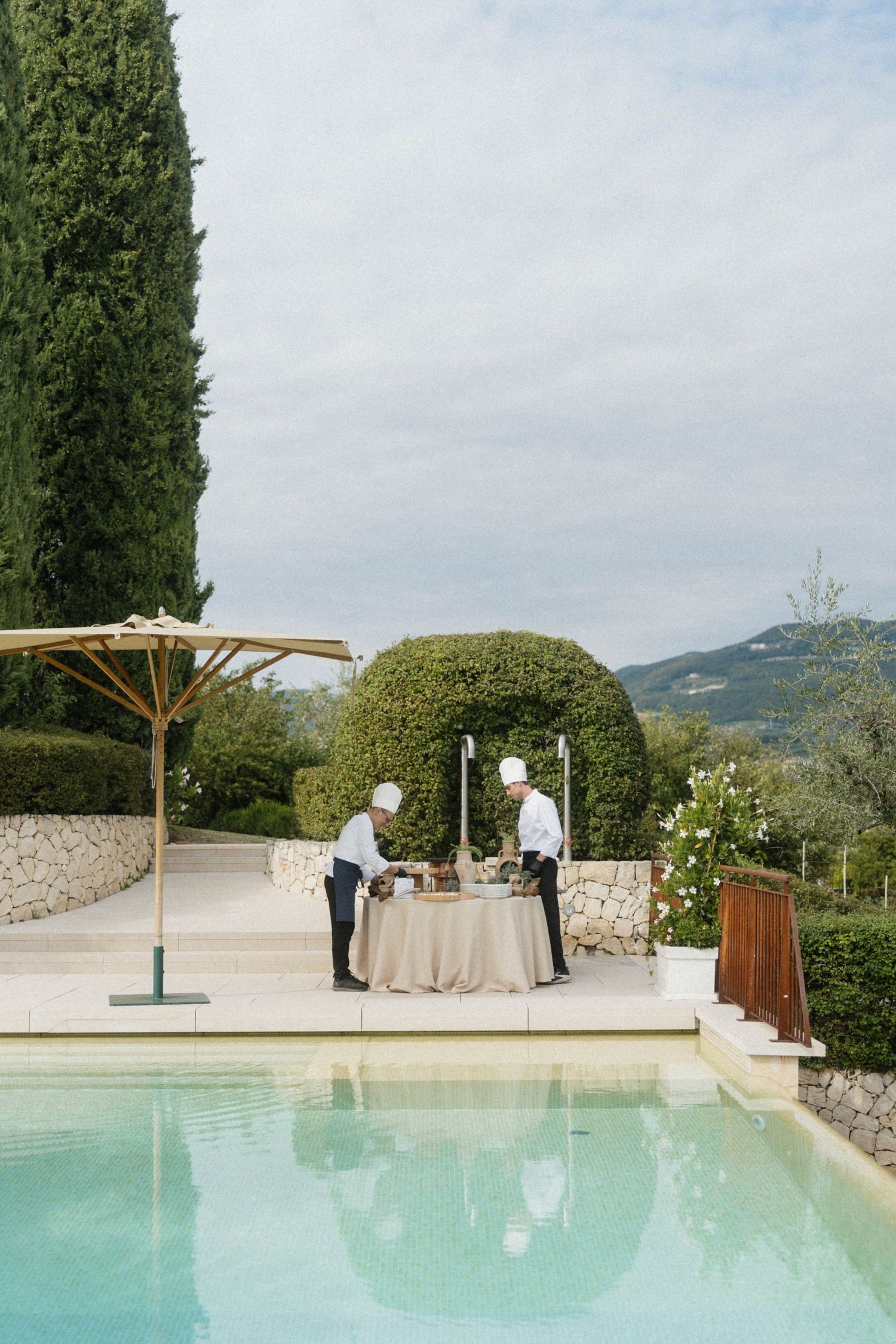 Two catering staff prepare a poolside table at an elegant villa with manicured hedges and mountain views.