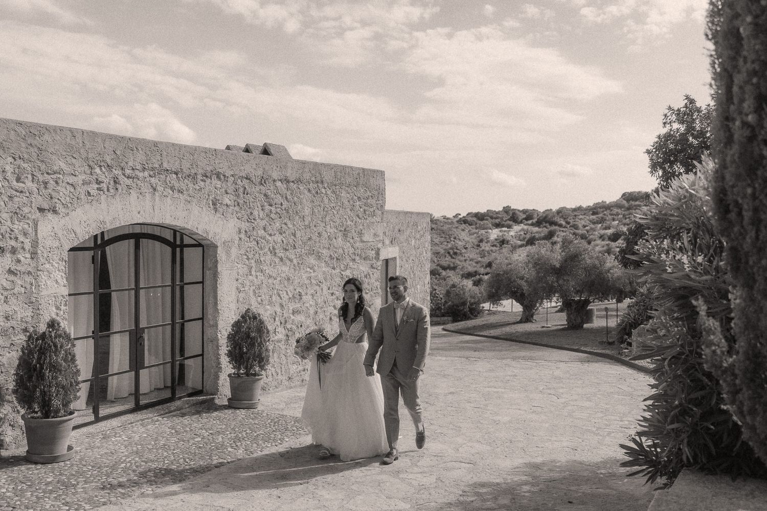 Black and white photo of a bride and groom walking outside a rustic stone venue surrounded by olive trees.