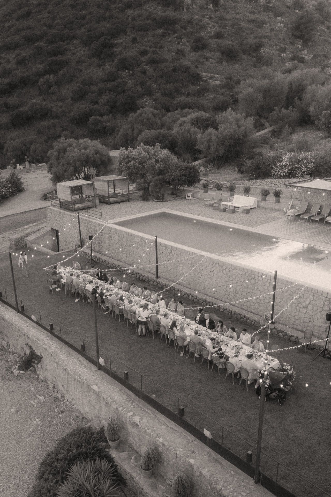 Aerial black and white photo of an outdoor wedding reception dinner beside a pool, surrounded by lush greenery.