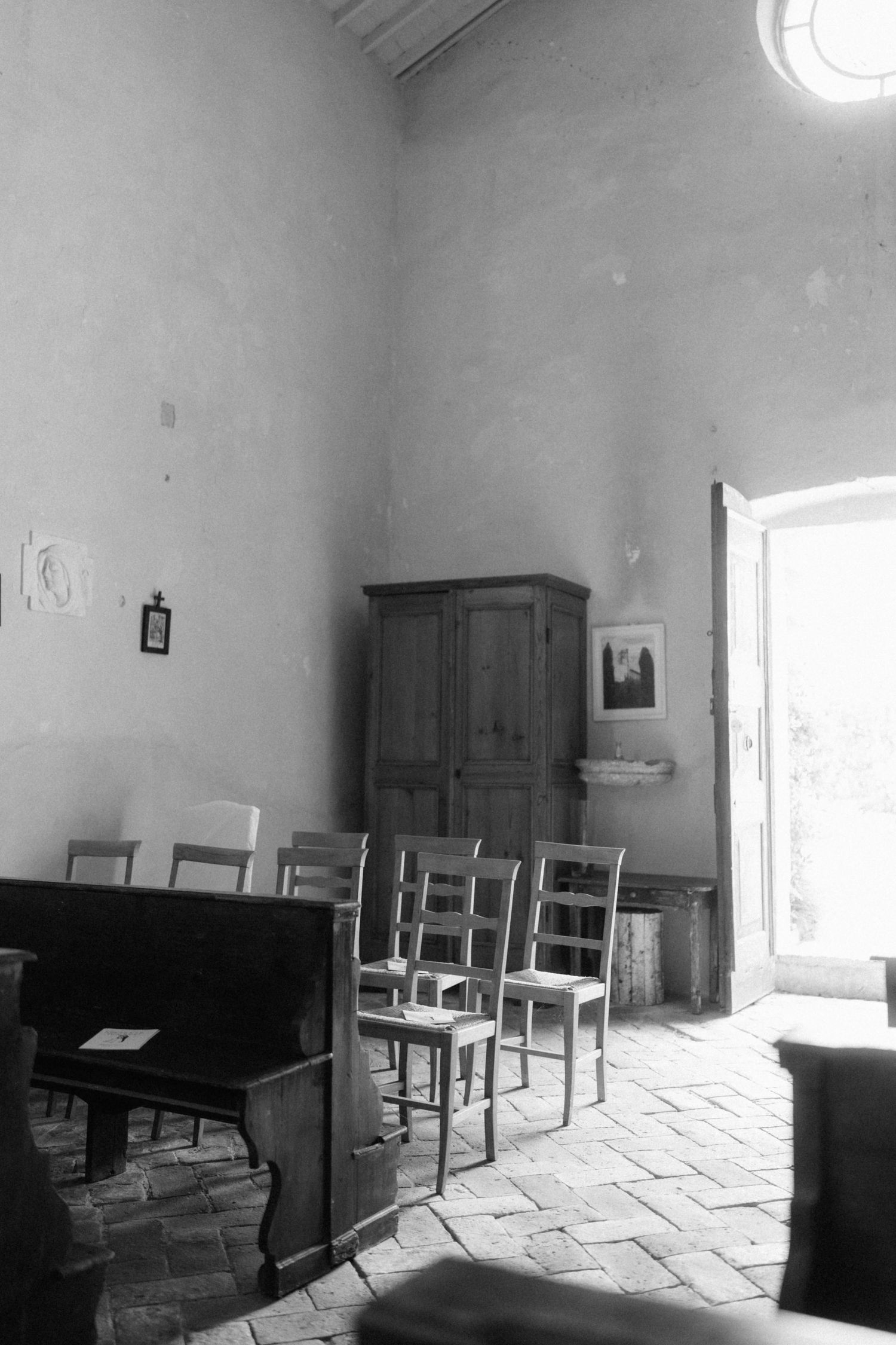 Black and white photo of a rustic room with wooden chairs, an antique armoire, and brick floors in an old building.