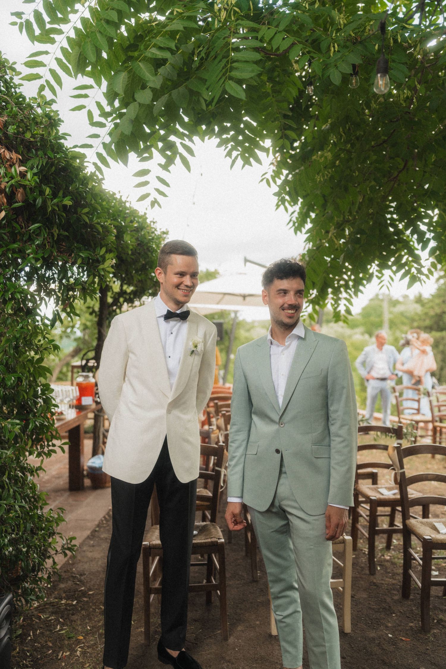 Two men in elegant suits walk together under a green leafy arch at an outdoor wedding.