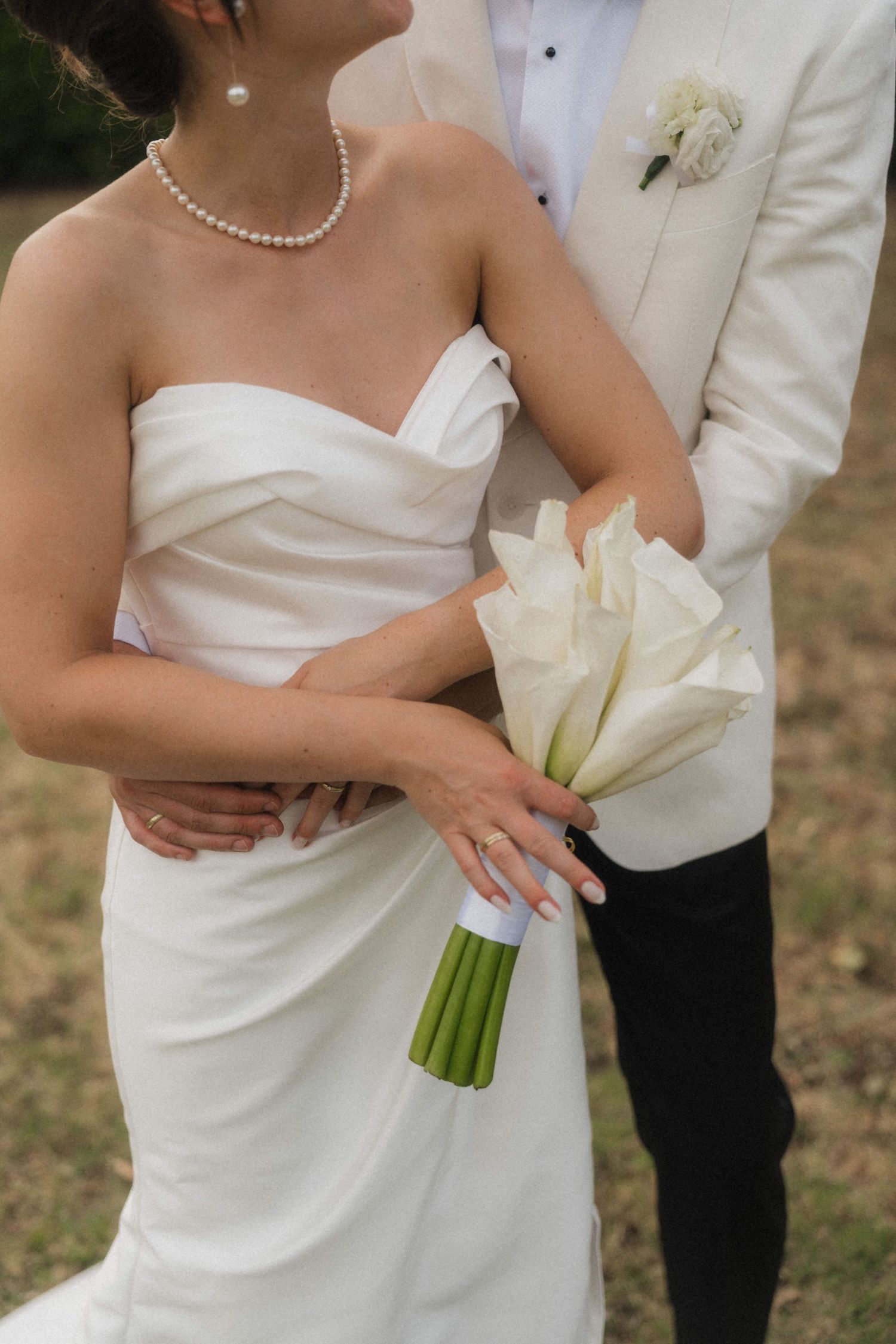 Bride in strapless white gown holding calla lily bouquet with groom in white tuxedo jacket embracing her outdoors.