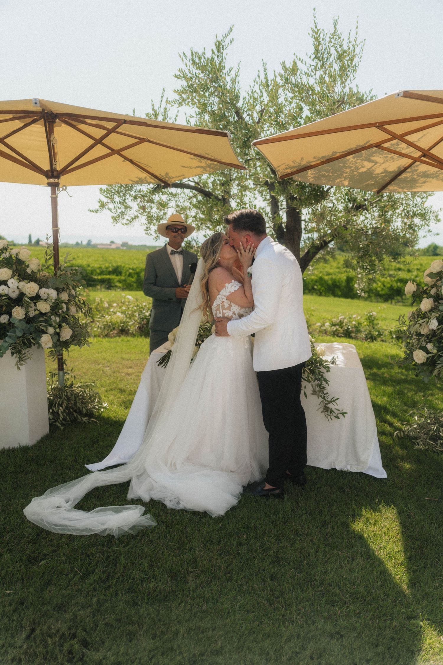 Bride and groom share first kiss at outdoor wedding ceremony surrounded by white floral arrangements.