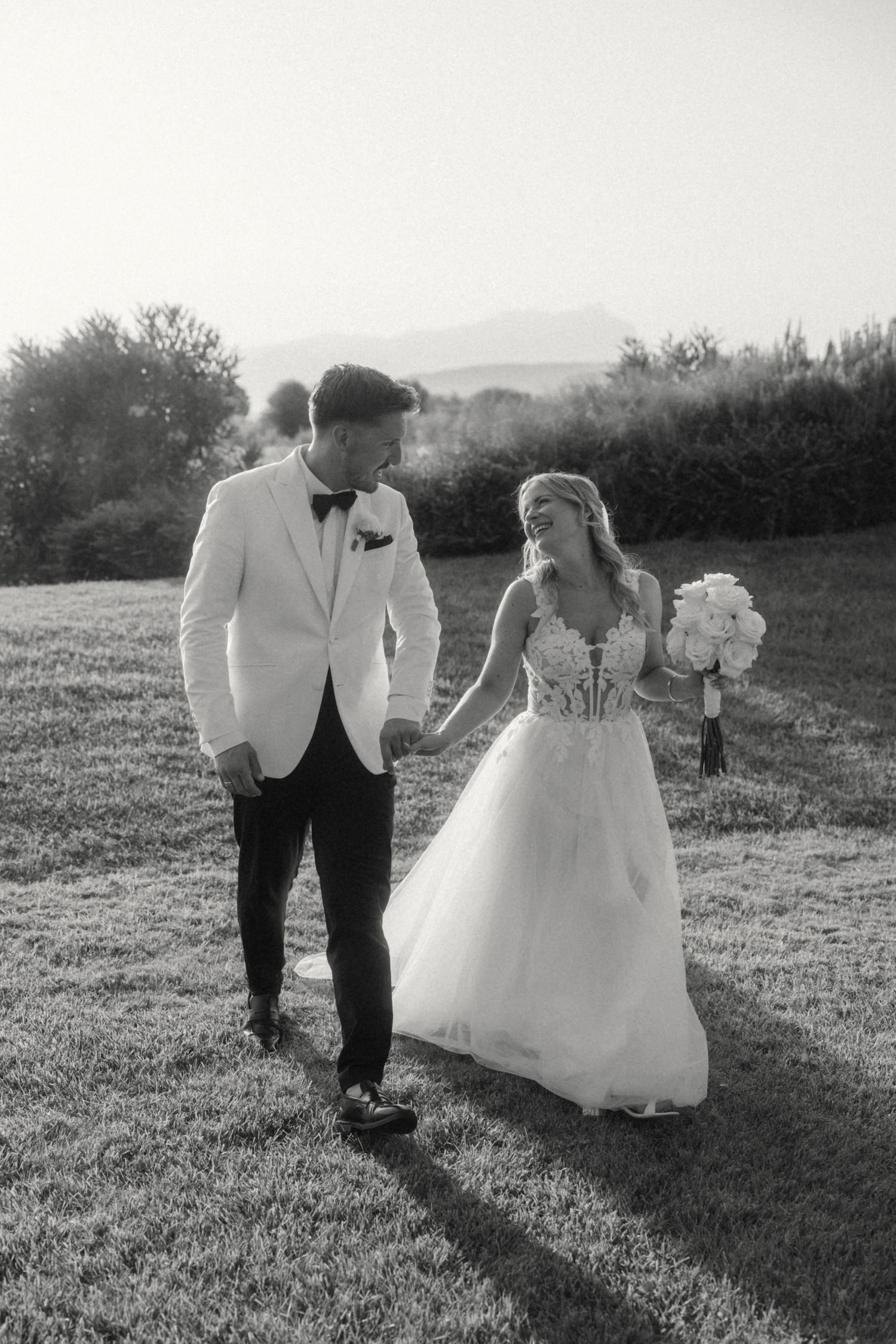 Black and white photo of a bride and groom holding hands while walking across a grassy field at sunset.
