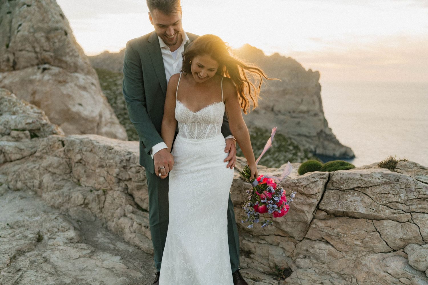 Bride and groom walk on rocky cliffs at sunset, bride holding vibrant bouquet in white gown.