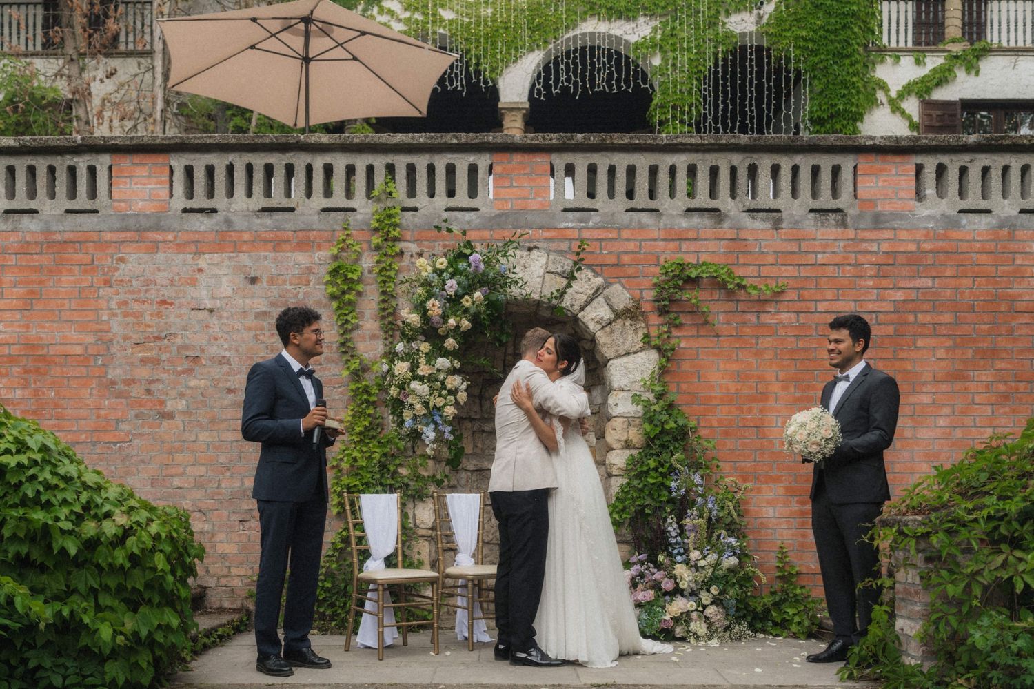 Outdoor wedding ceremony at brick wall garden with floral arch, couple embracing before officiant.