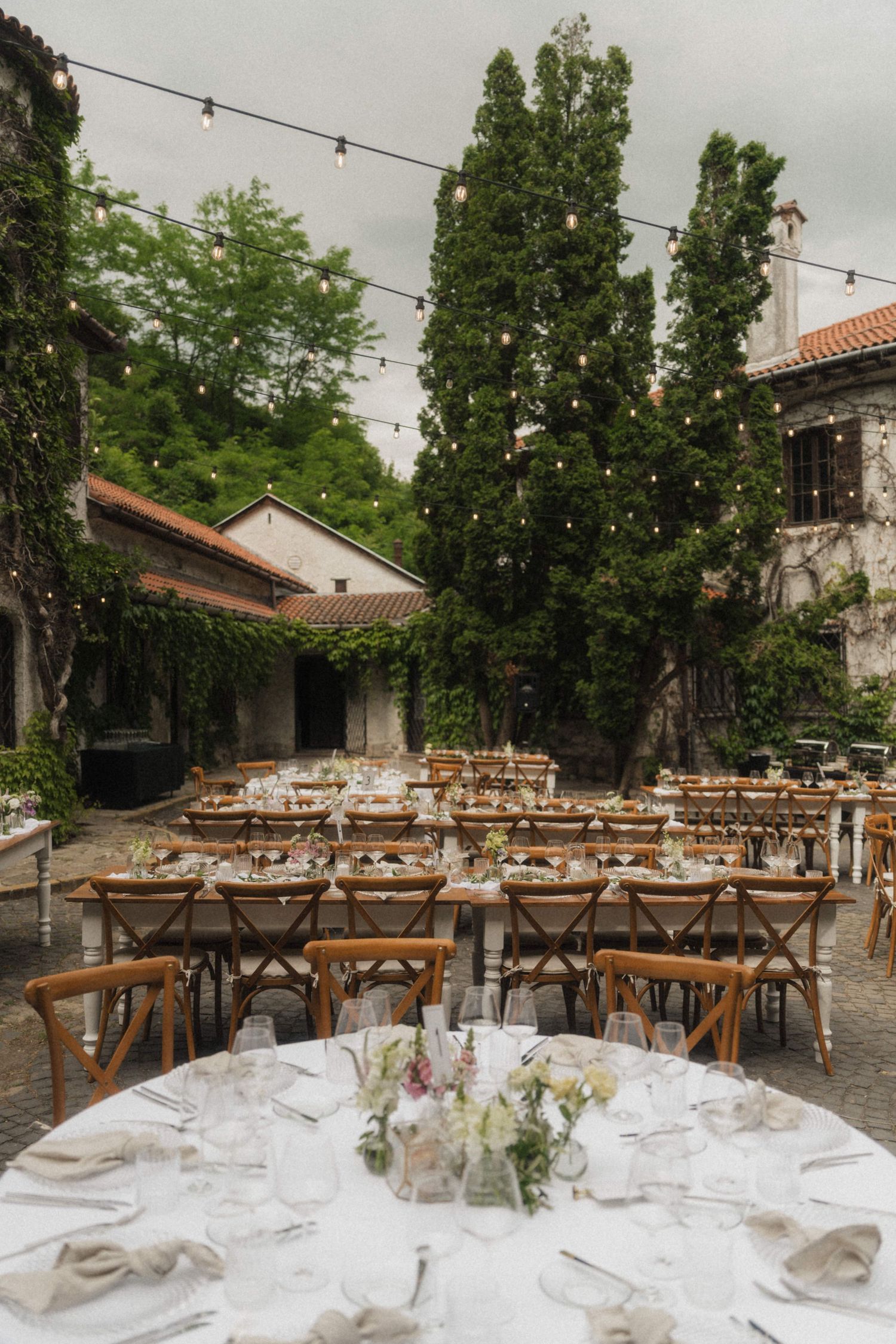 Outdoor wedding reception courtyard with wooden chairs, long tables, and string lights at dusk.