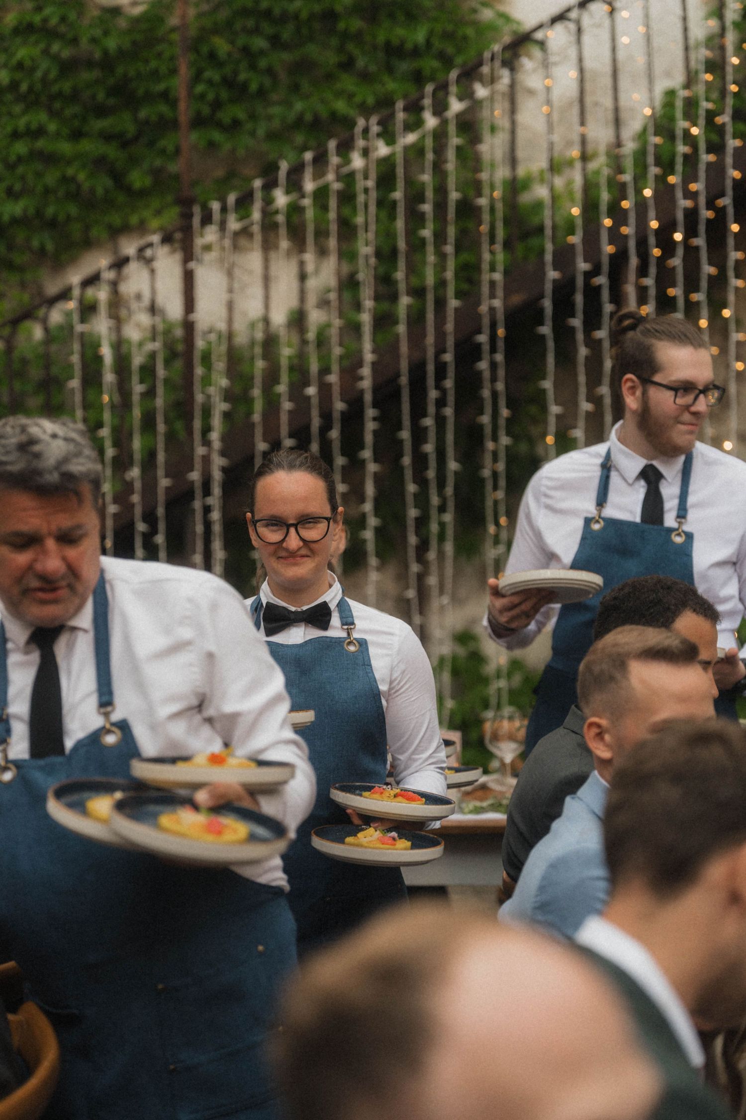 Smiling waitstaff in blue aprons serving dishes to guests at an outdoor wedding reception with fairy lights.