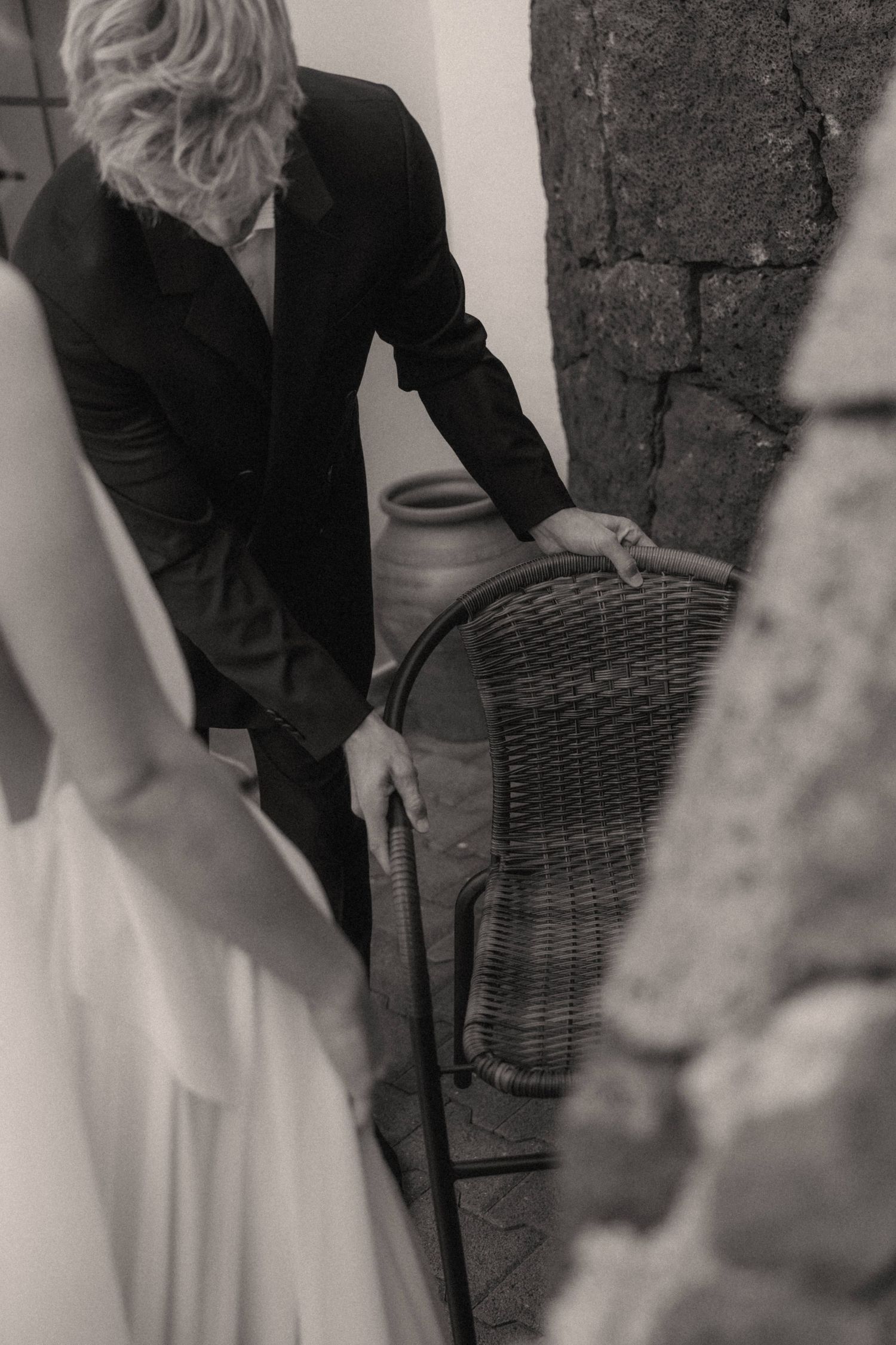 Black and white photo of a groom in a suit pulling out a wicker chair for the bride in a white dress.