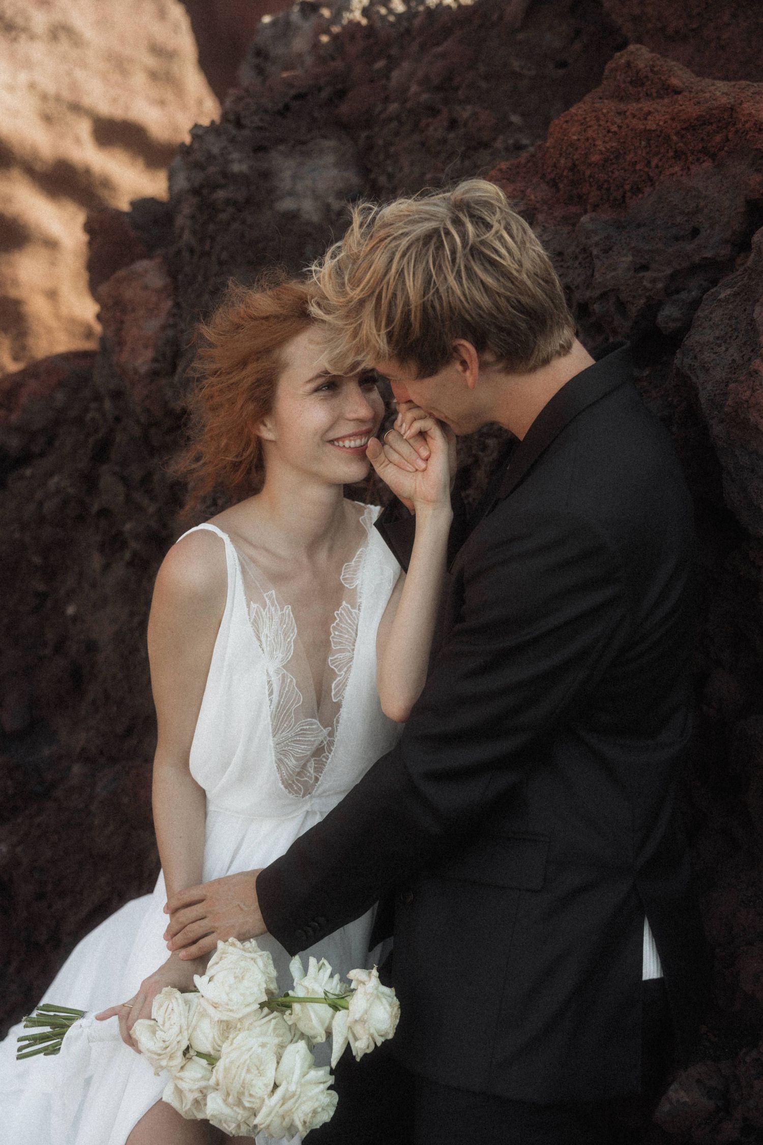 Smiling bride in white lace dress holds bouquet as groom whispers to her by red volcanic rock.