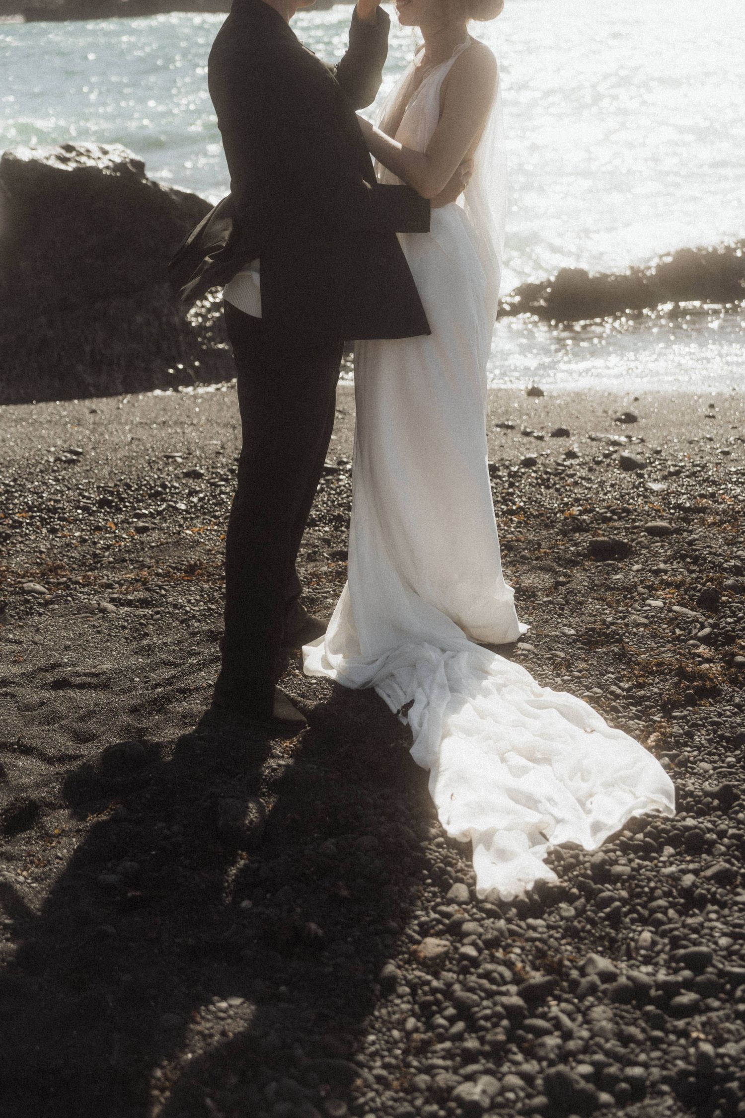Wedding couple embracing on rocky black sand beach with ocean waves in background.