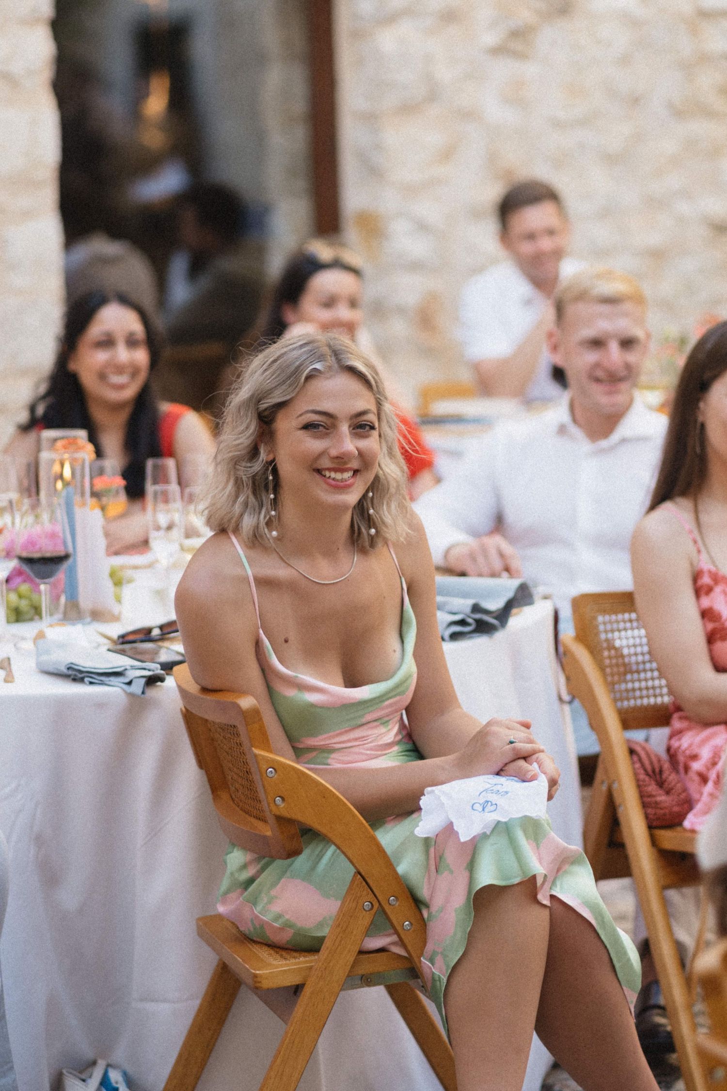 Smiling blonde wedding guest in a pink and green dress seated at an outdoor reception table.