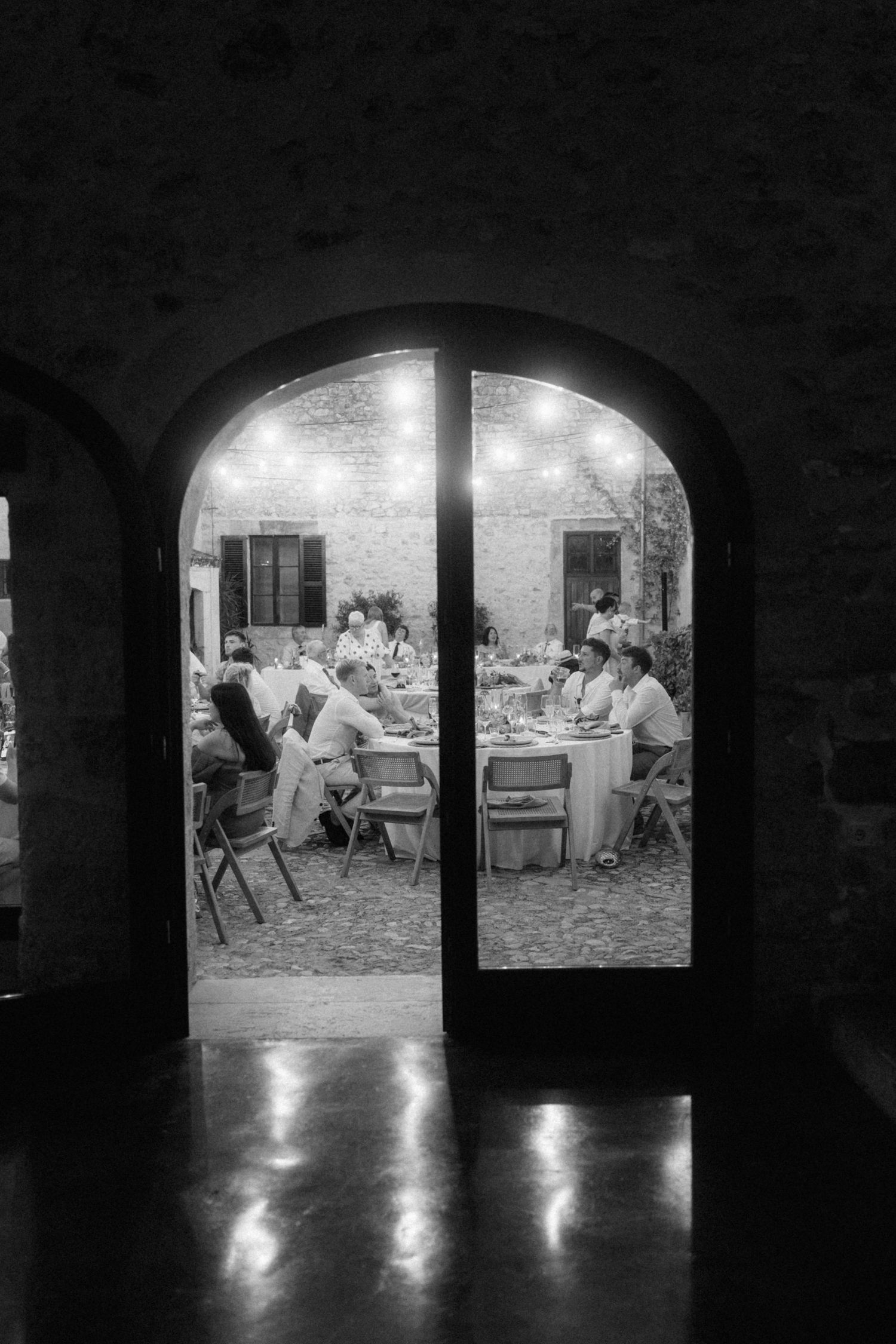 Black and white view through arched doorway of wedding guests dining under string lights at night.
