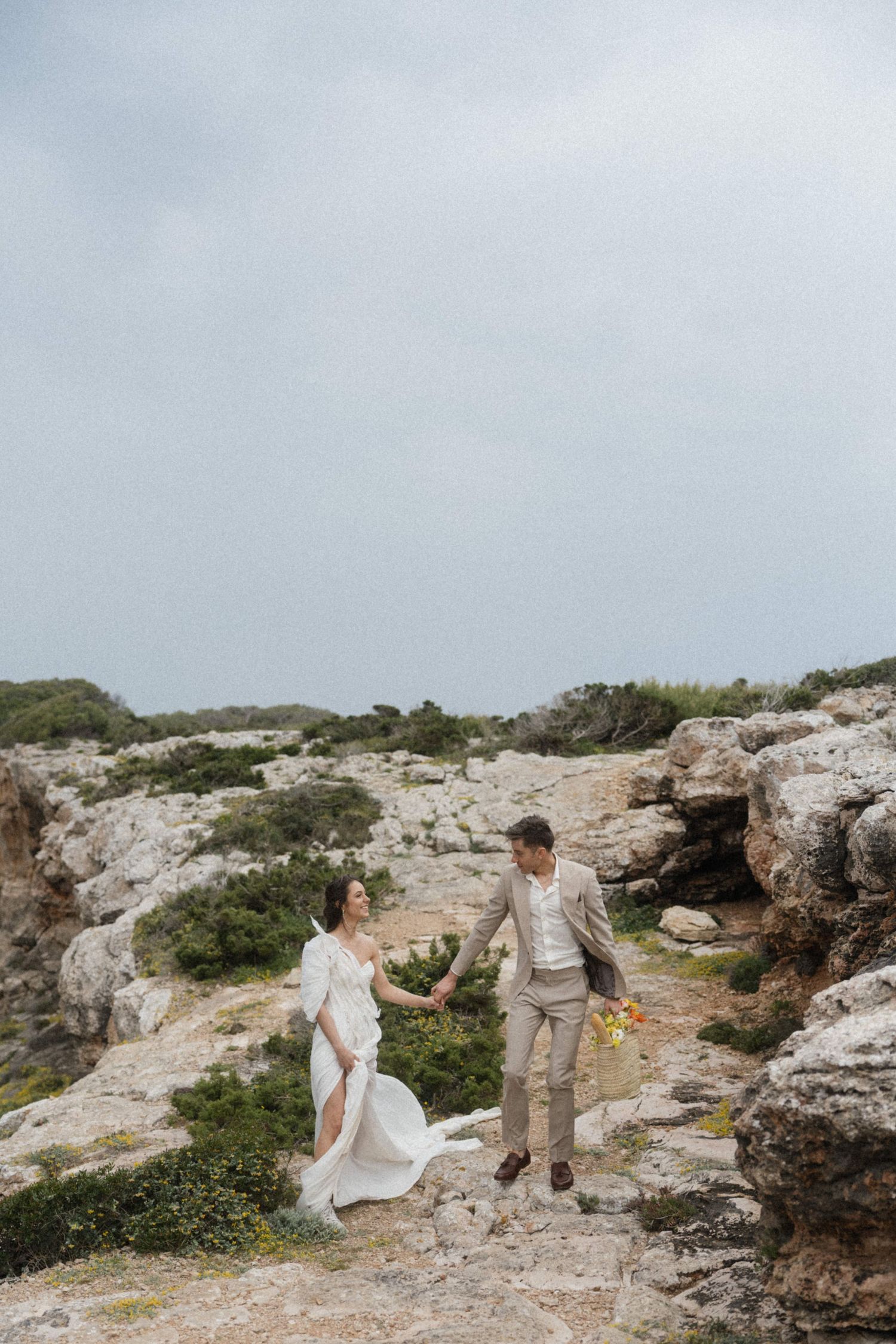 Wedding couple holding hands walking on rocky coastal landscape under overcast sky.