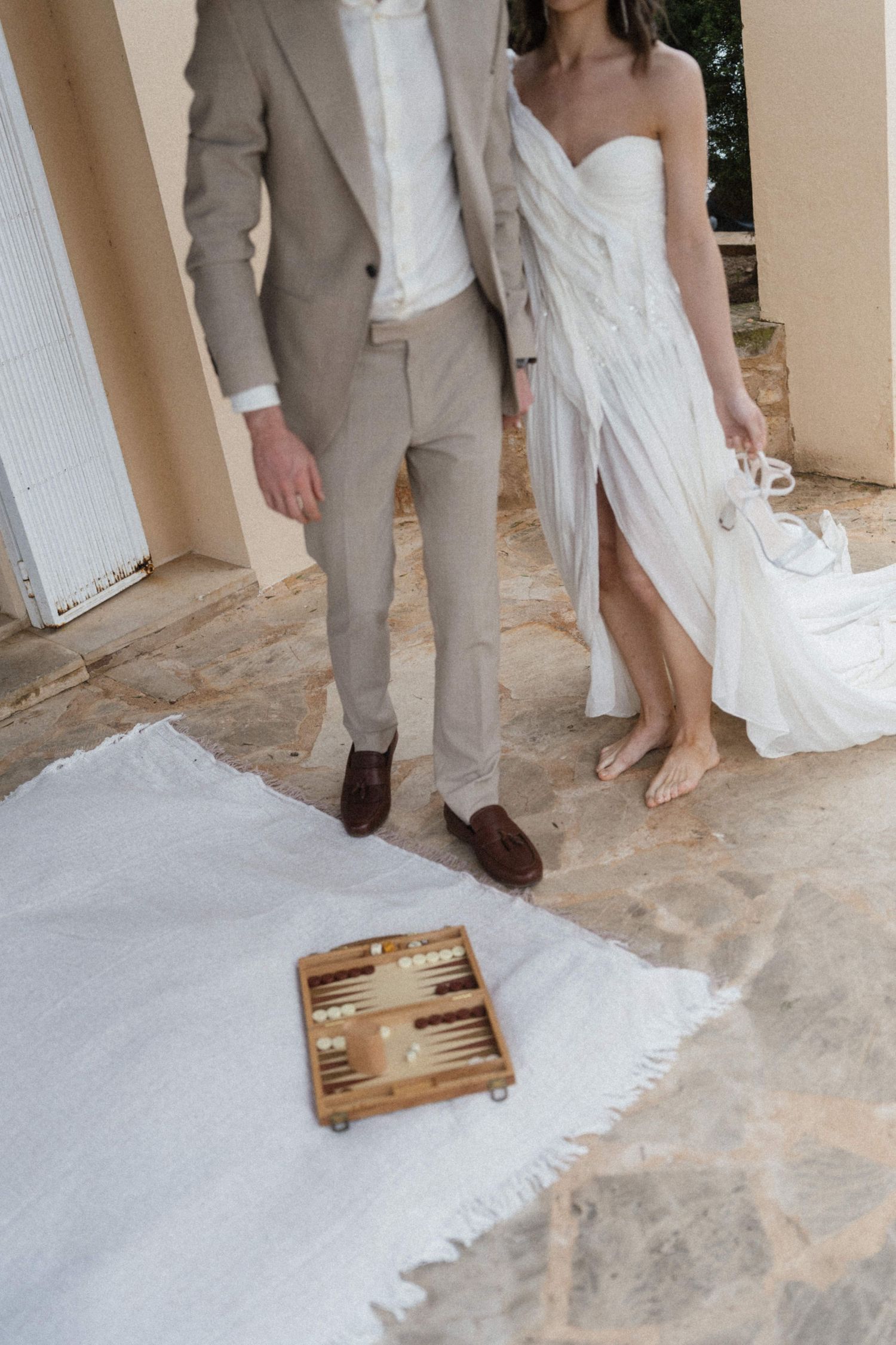 Couple in wedding attire standing on stone patio with backgammon board on white blanket.