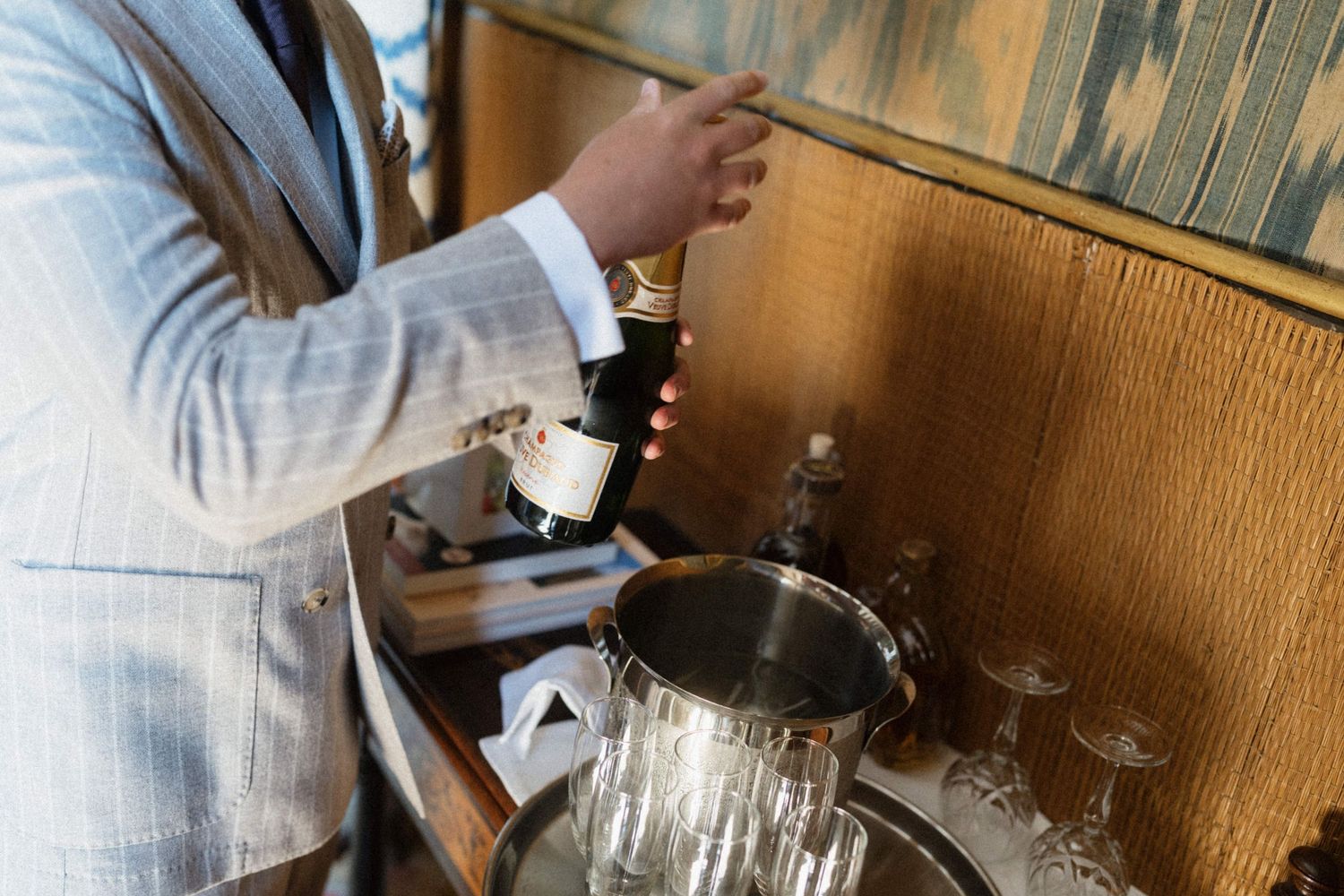 Elegant man in gray suit pouring champagne into silver ice bucket at a sophisticated bar setting.