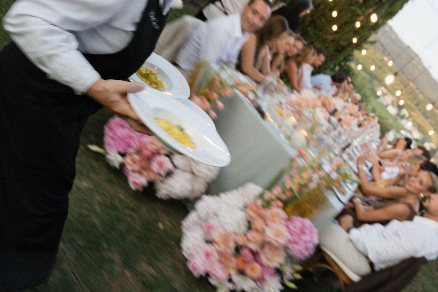 Waiter serves food at a floral-decorated outdoor wedding reception dinner table at dusk.