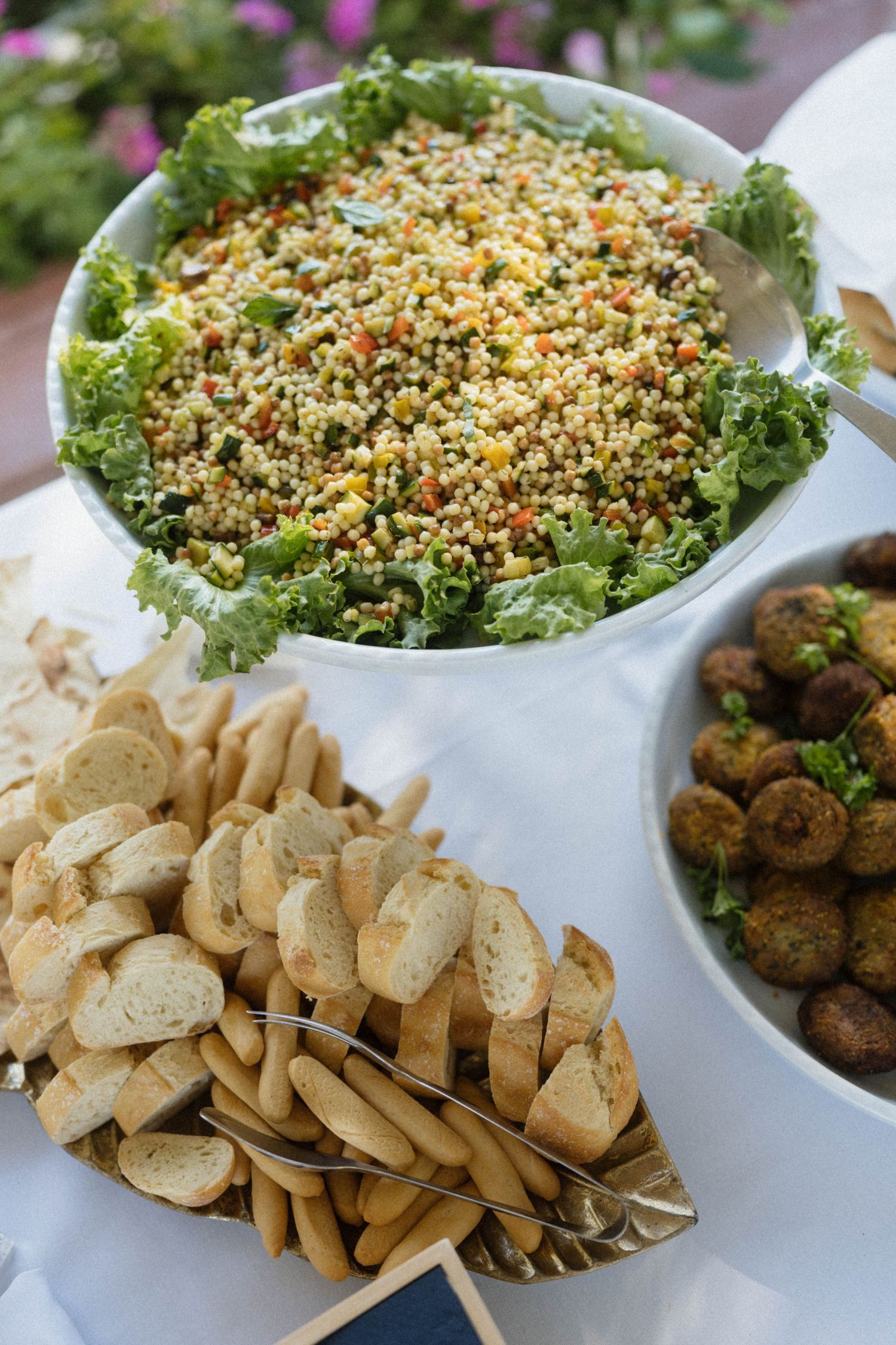 Buffet spread featuring quinoa salad on kale, sliced toasted bread, and roasted falafel balls in white serving bowls.