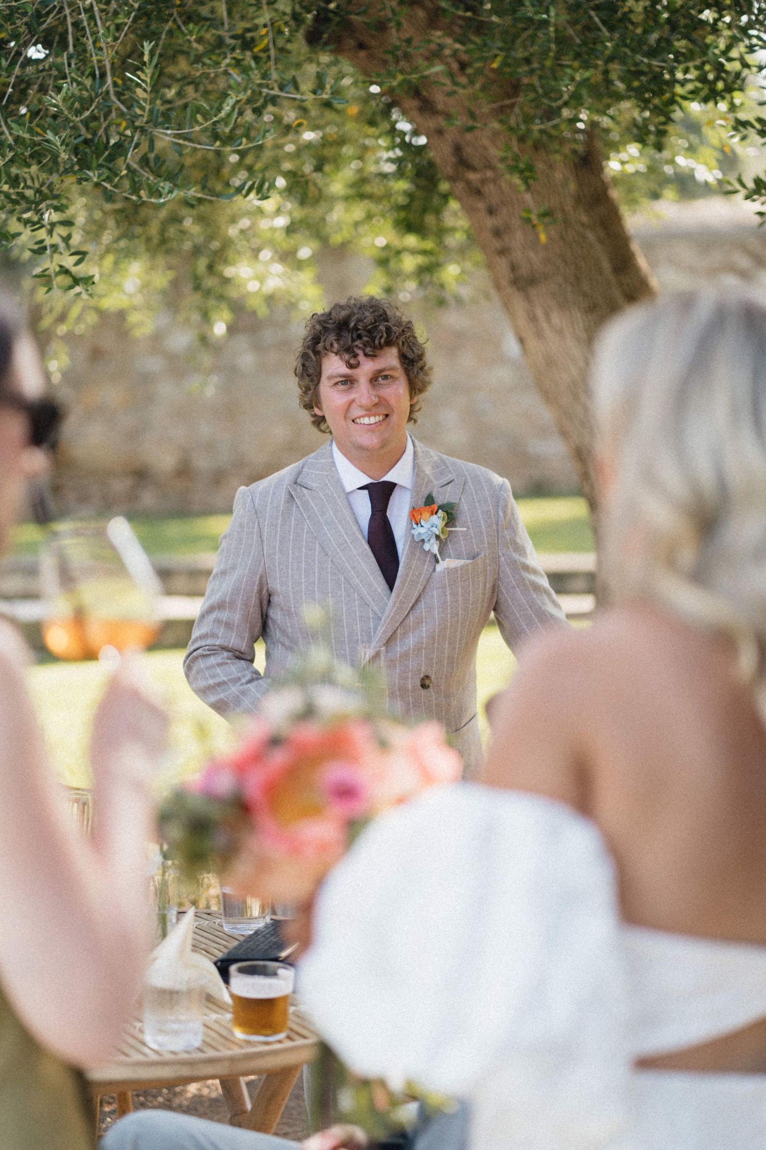 Smiling groom in striped suit with boutonniere stands outdoors facing bride holding colorful wedding bouquet.