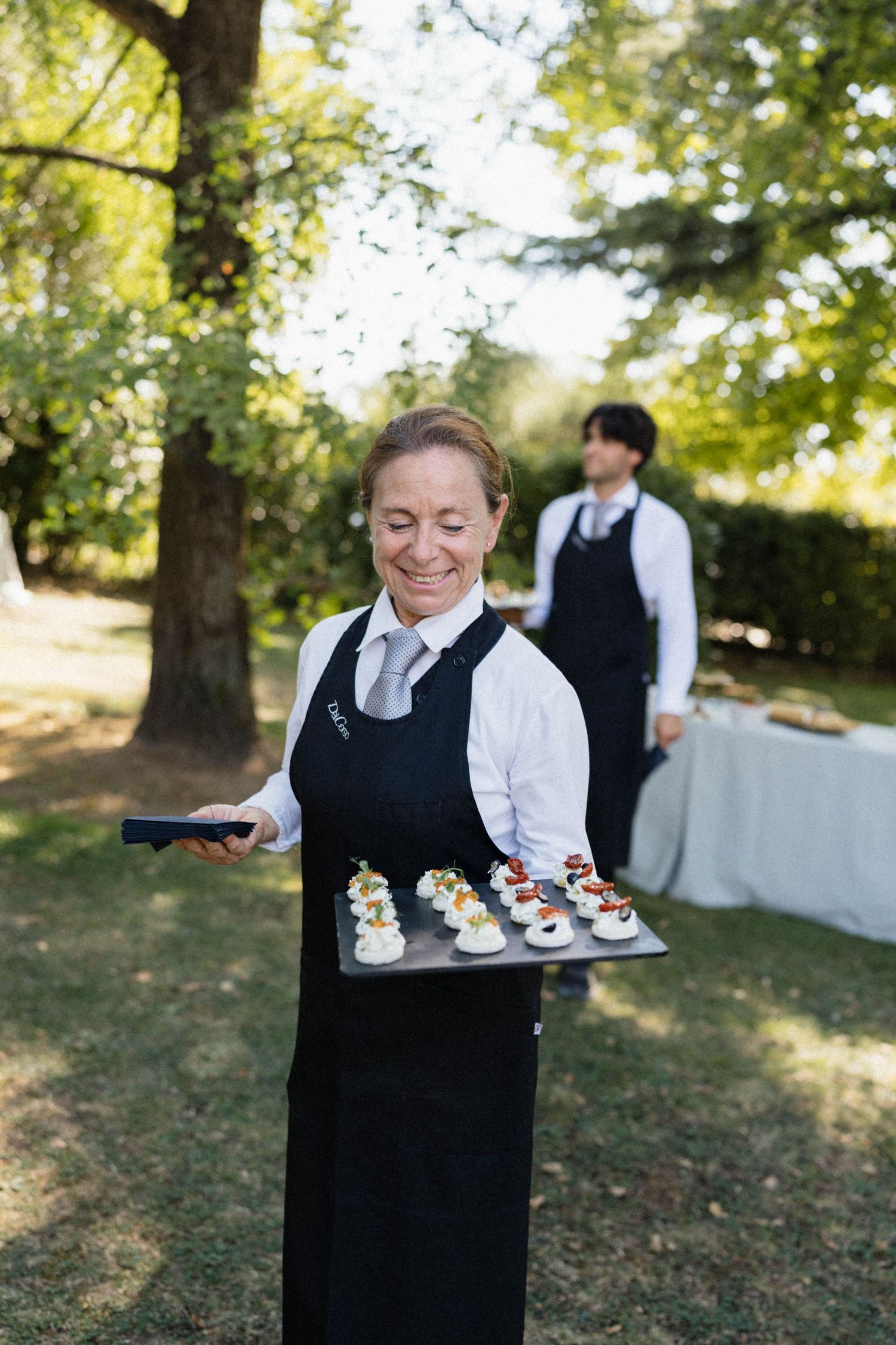 Smiling female caterer in black apron carries tray of elegant appetizers at outdoor garden event.
