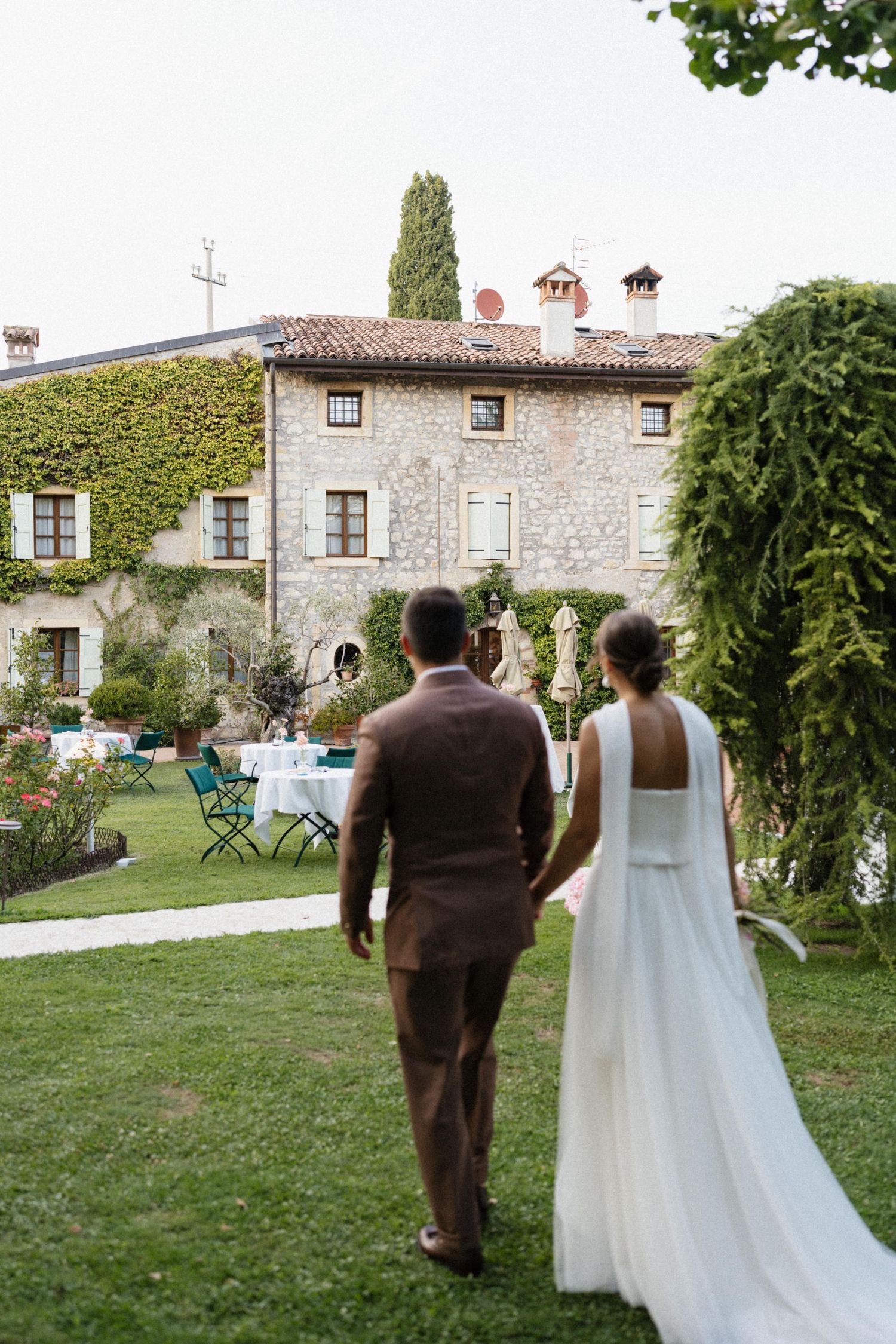 Bride and groom walking hand in hand toward ivy-covered stone villa with garden reception setup.