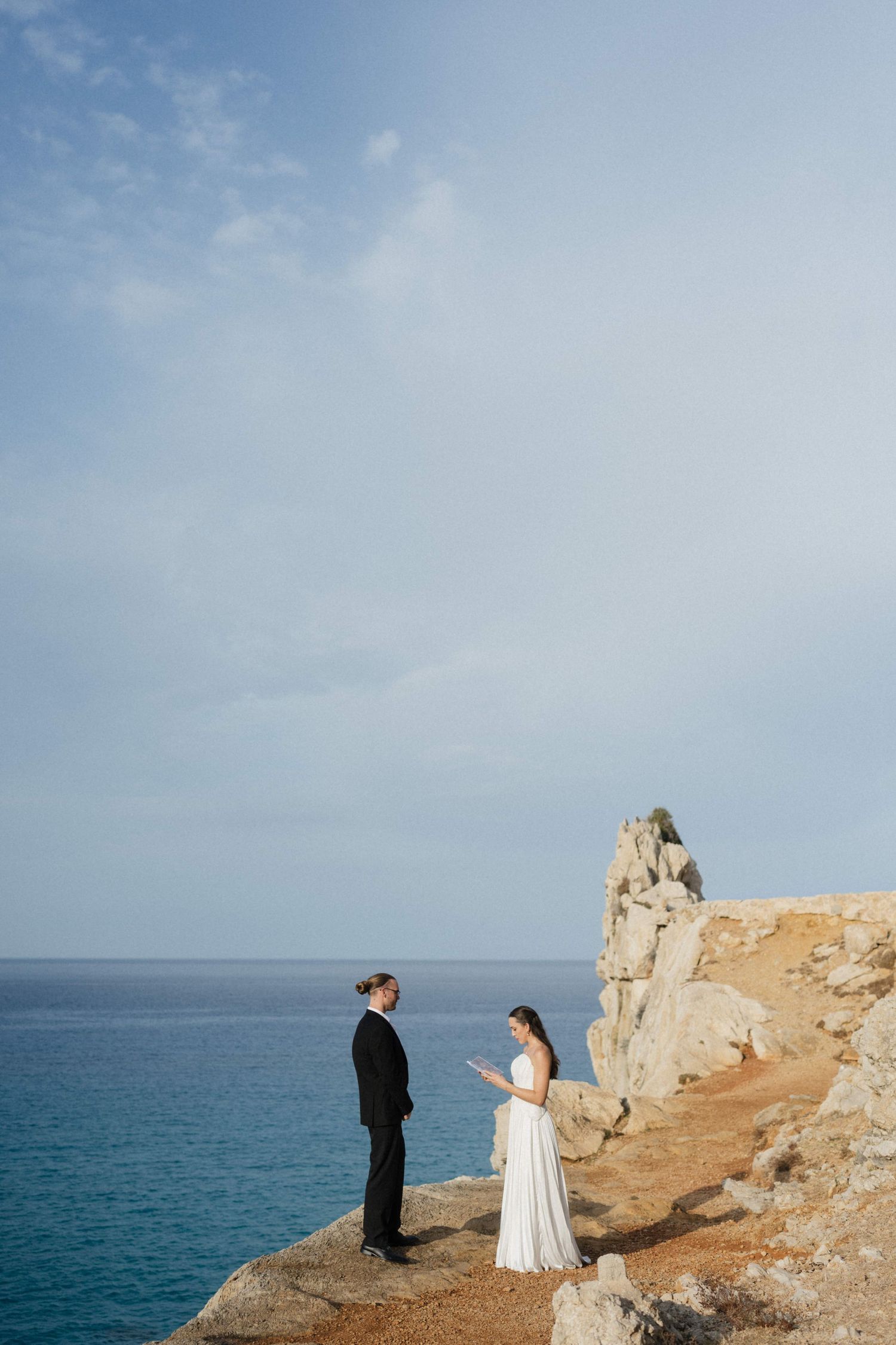 Bride in a white gown and groom in a black suit stand facing each other on a rocky seaside cliff above the turquoise sea.