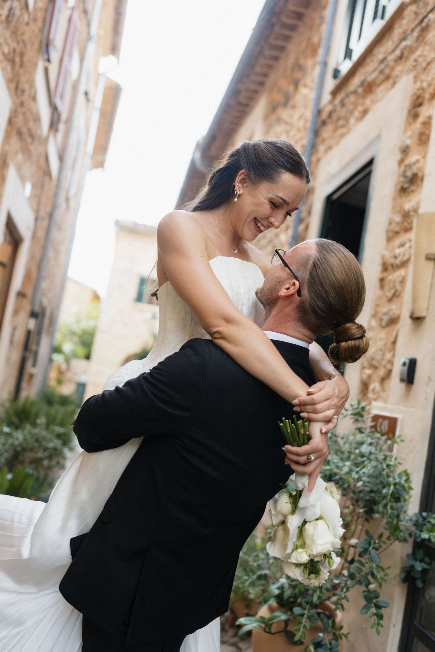 Smiling bride in a white pleated gown is lifted by her groom in a black suit holding a bouquet on a charming stone street.