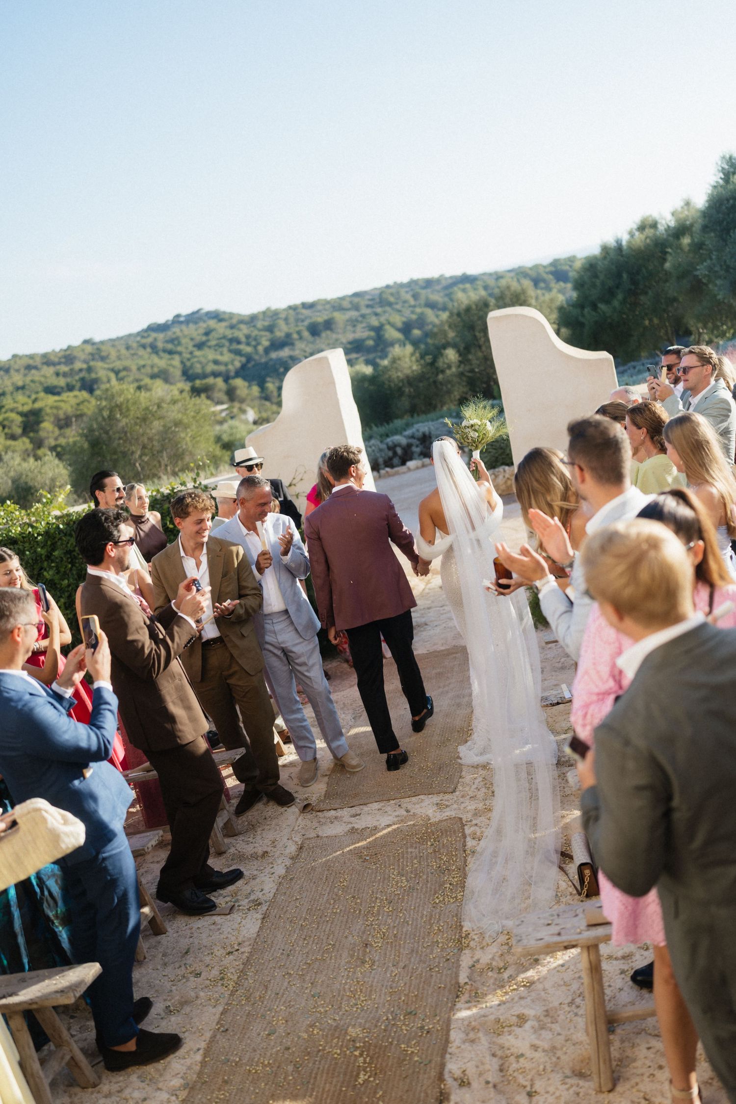 Wedding guests clap and cheer as the groom leads the veiled bride down a stone path after the ceremony.