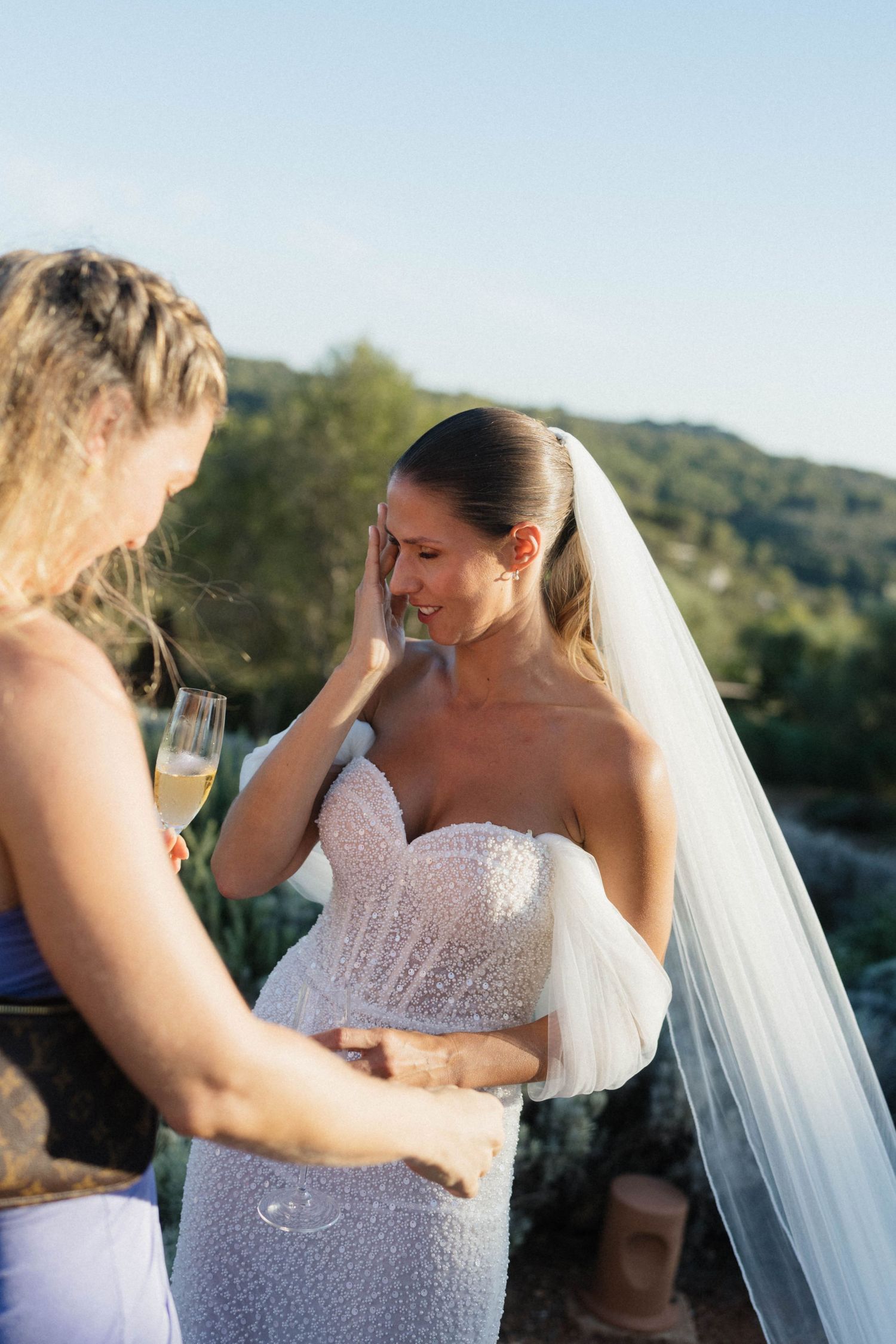 A emotional bride in a beaded strapless wedding gown and long veil wipes a tear as a friend holds a champagne glass.
