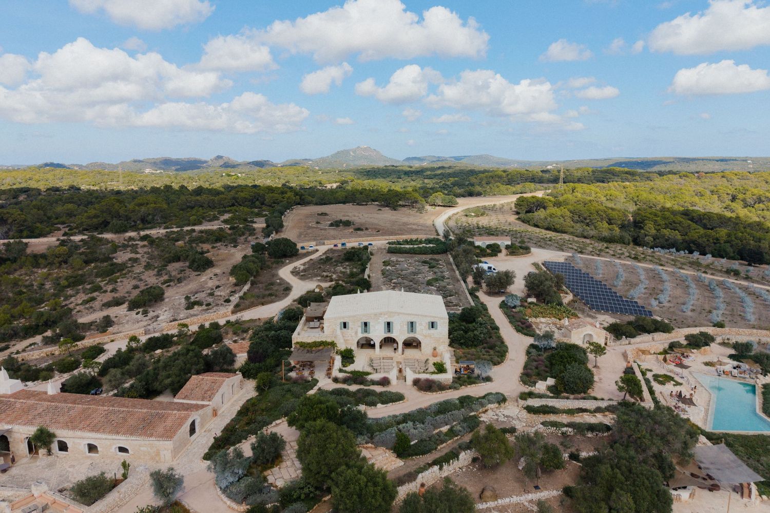 Aerial view of a whitewashed Mediterranean farmhouse with a swimming pool, terraced gardens and rolling countryside under a blue sky.