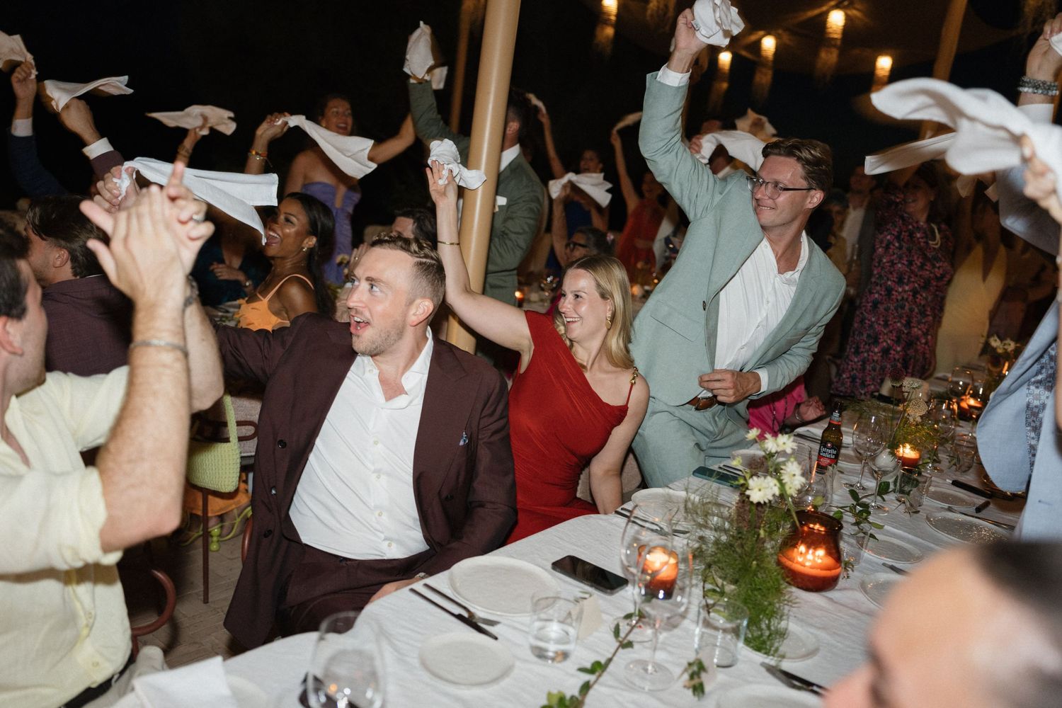 Wedding guests cheering and waving white napkins in the air at a candlelit reception dinner table.