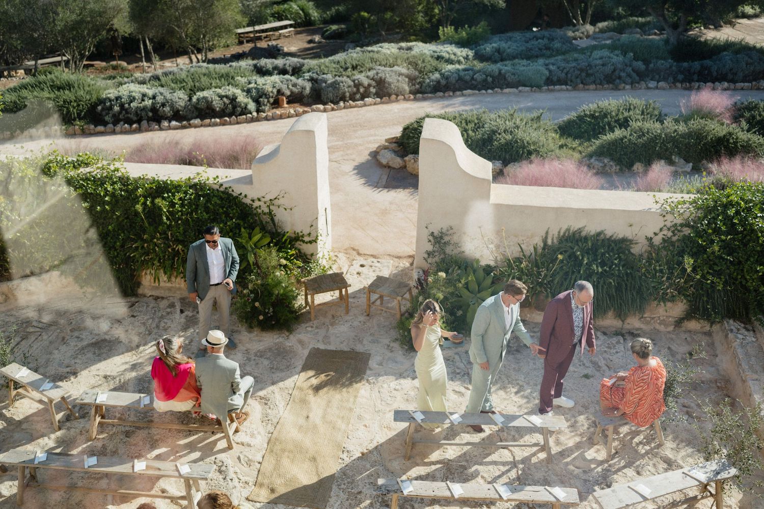 Wedding guests gather along a sandy path lined with whitewashed walls and Mediterranean shrubs before an outdoor ceremony.