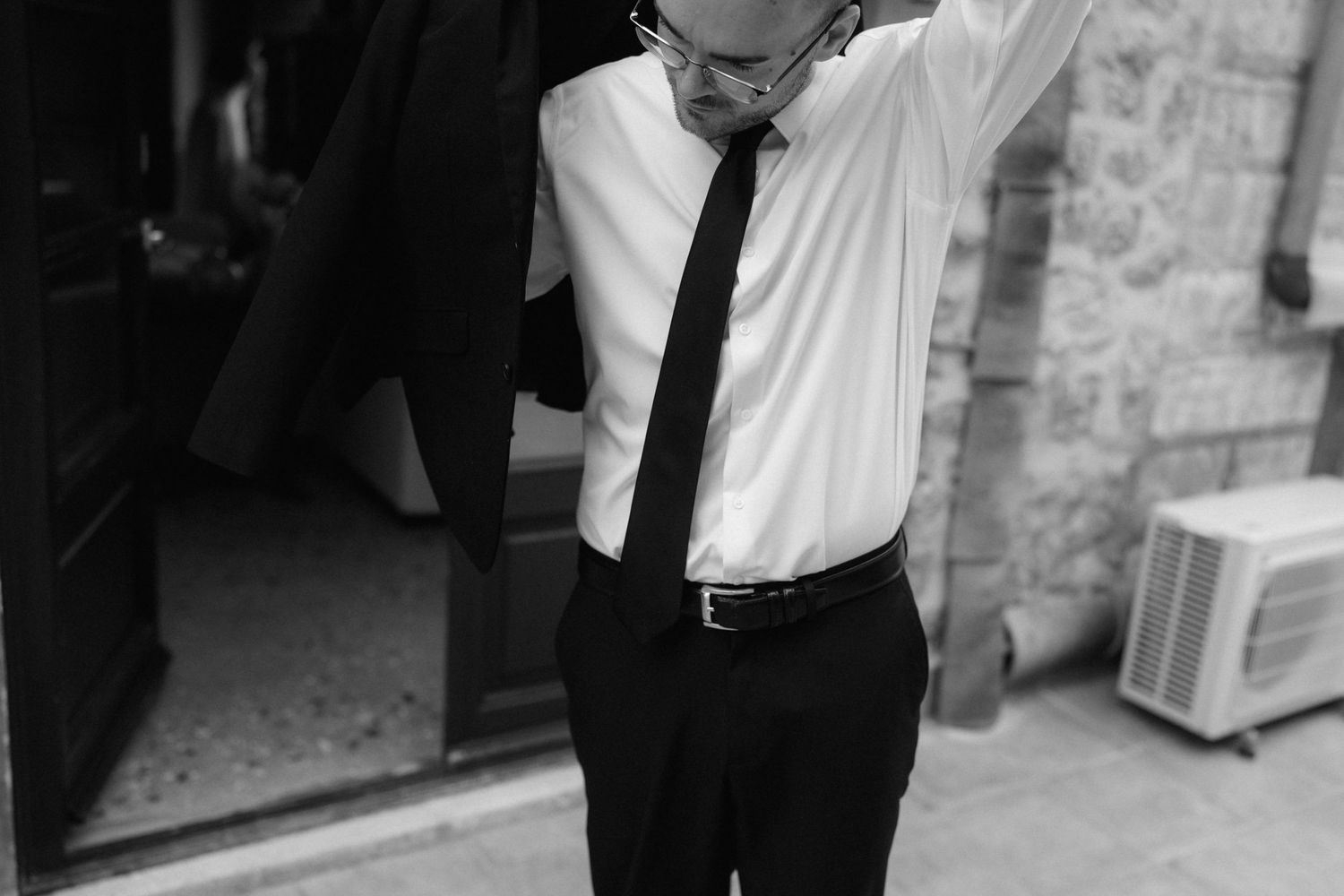 Black and white photo of a man in glasses, white shirt and tie removing his jacket outside a stone building doorway.