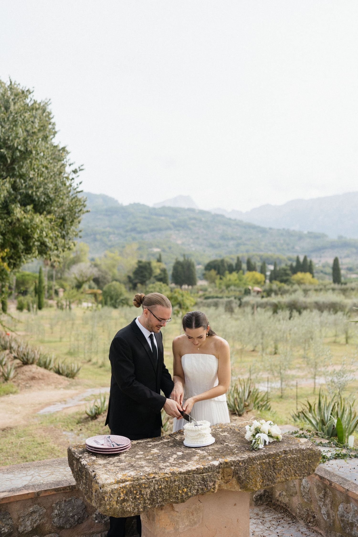 Bride and groom cut their wedding cake together on a rustic stone table overlooking olive groves and mountains.