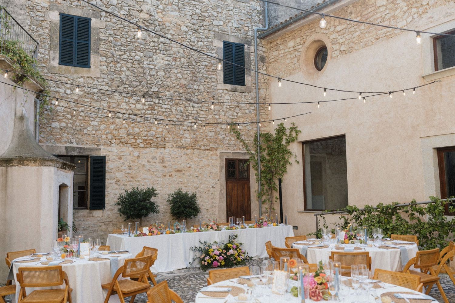 Elegant outdoor wedding reception in a rustic stone courtyard with string lights, round tables, and floral centerpieces.