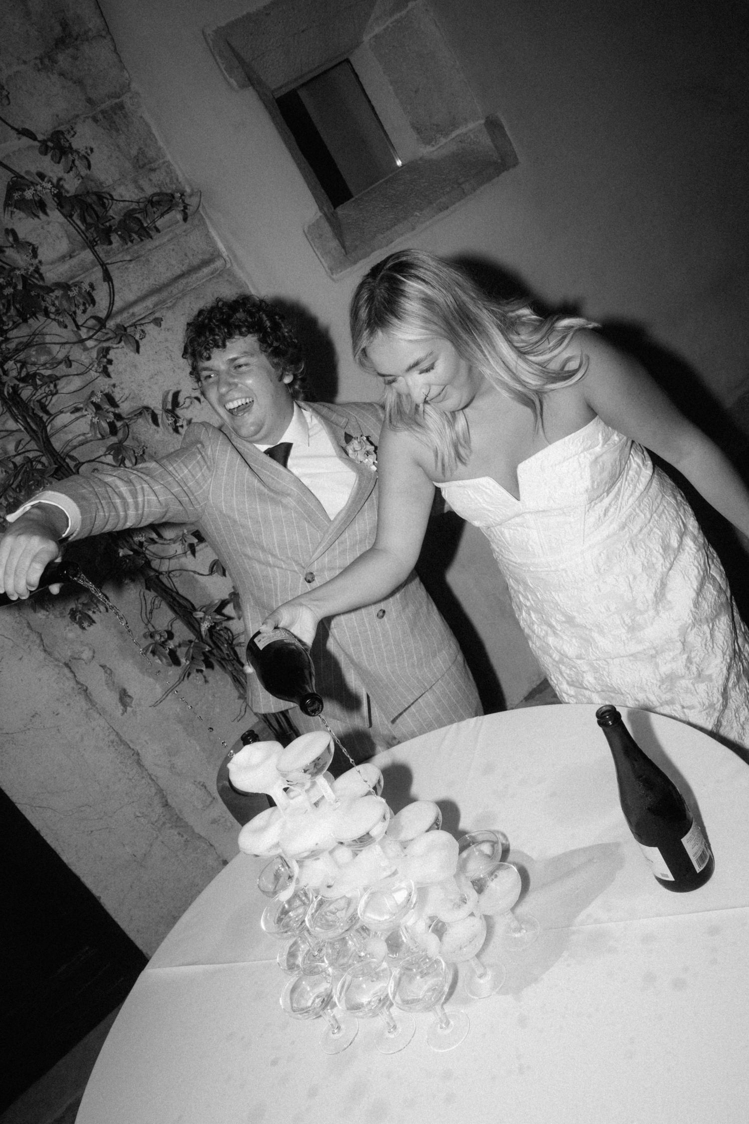 Black and white photo of a bride and groom laughing while pouring champagne tower at their wedding reception.
