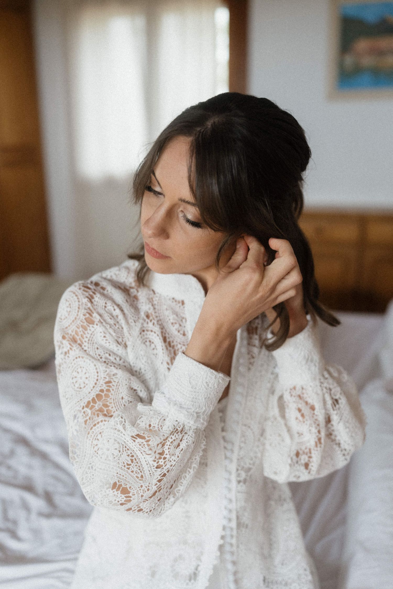 Elegant bride in white lace robe adjusting her hair while sitting on a bed near a window.