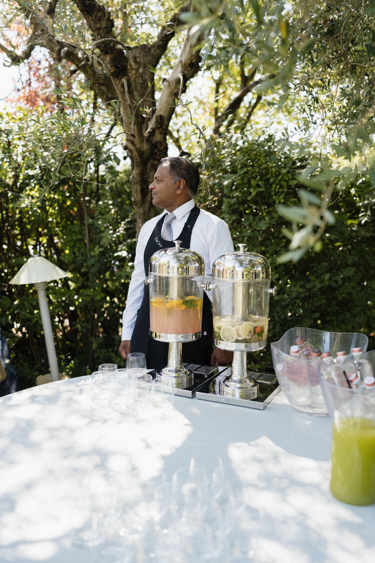 Bartender stands behind beverage dispensers filled with colorful drinks at an outdoor wedding reception.