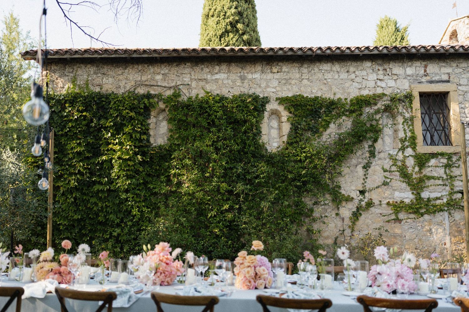 Elegant outdoor wedding reception table with pink floral centerpieces set against an ivy-covered stone villa in Tuscany.