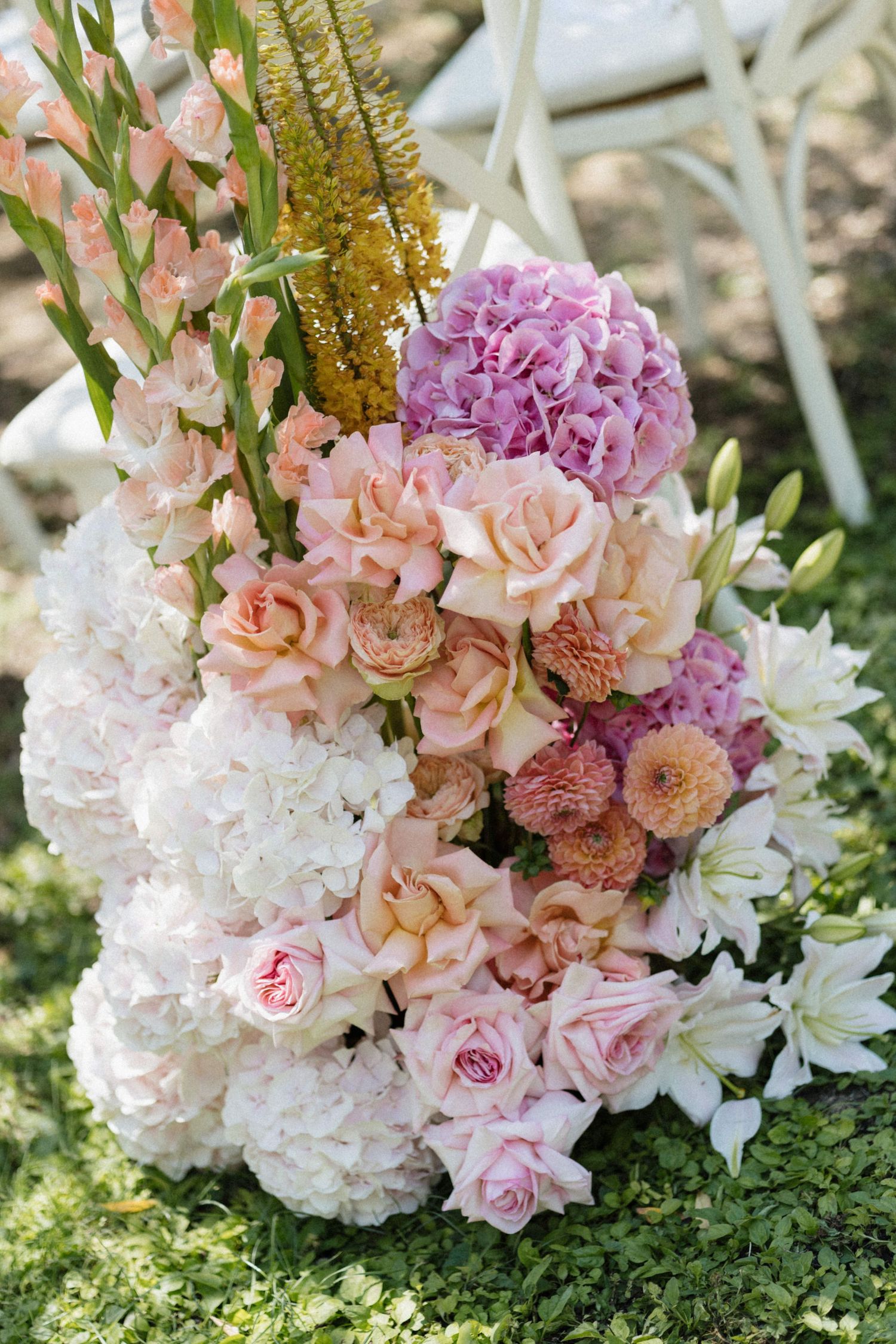 Elegant outdoor floral arrangement featuring pink roses, white hydrangeas, purple blooms, and golden accents for a wedding ceremony.